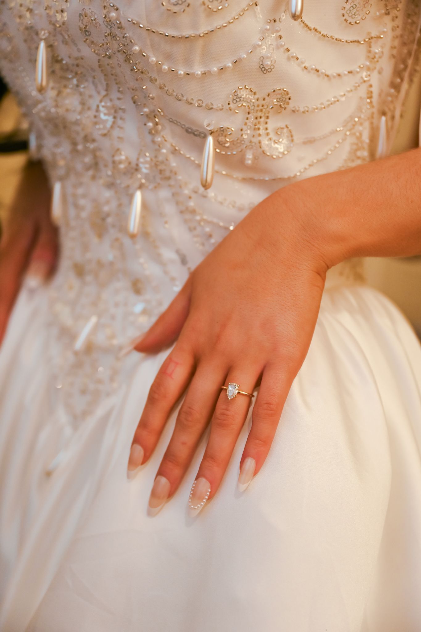 Close-up view of a hand wearing an engagement ring resting on white wedding dress fabric with beaded details.