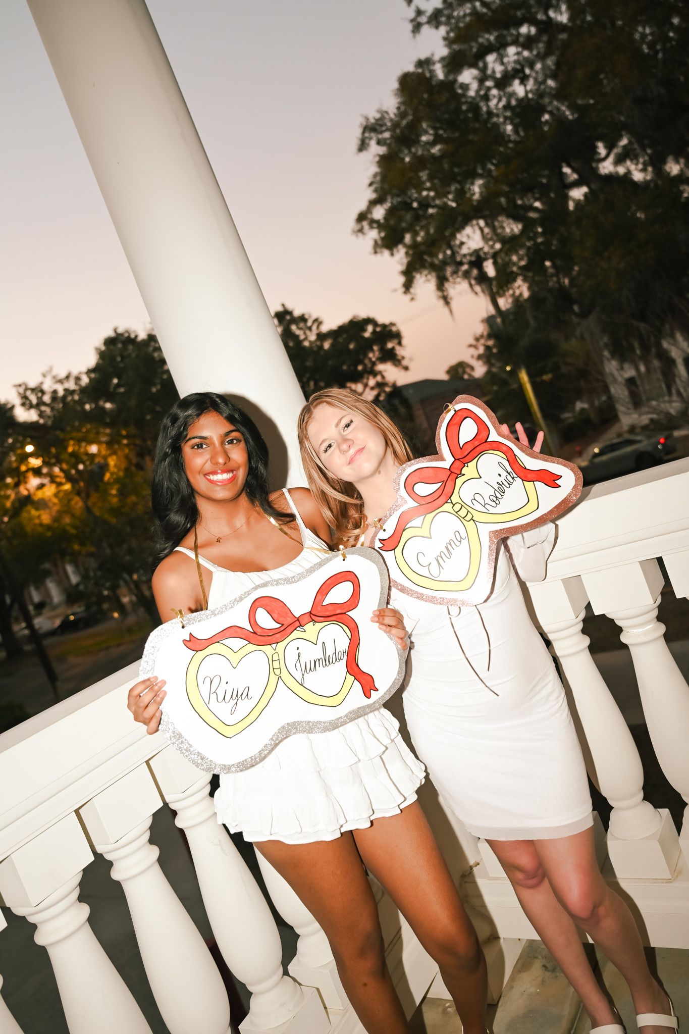 Two smiling women in white dresses hold decorative heart-shaped signs with red bows on a white balcony at dusk.