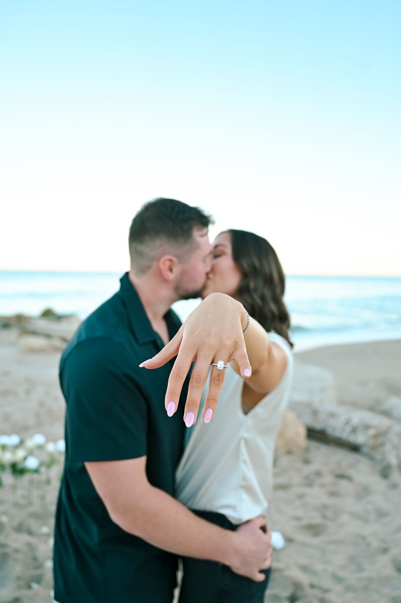 A couple embraces and shares a romantic moment on a sandy beach at sunset with ocean waves in the background.
