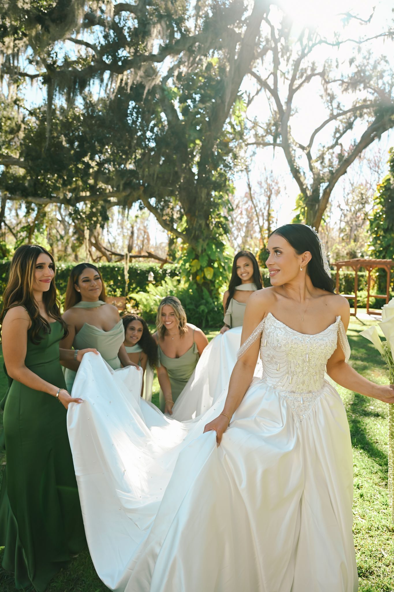 Bride in white gown with bridesmaids in sage green dresses holding her train outdoors under Spanish moss trees.