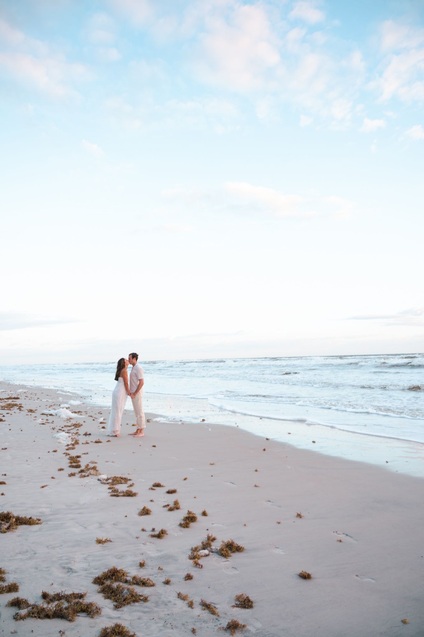 Couple embracing on a serene white sand beach at dusk with gentle ocean waves and a pastel blue sky.