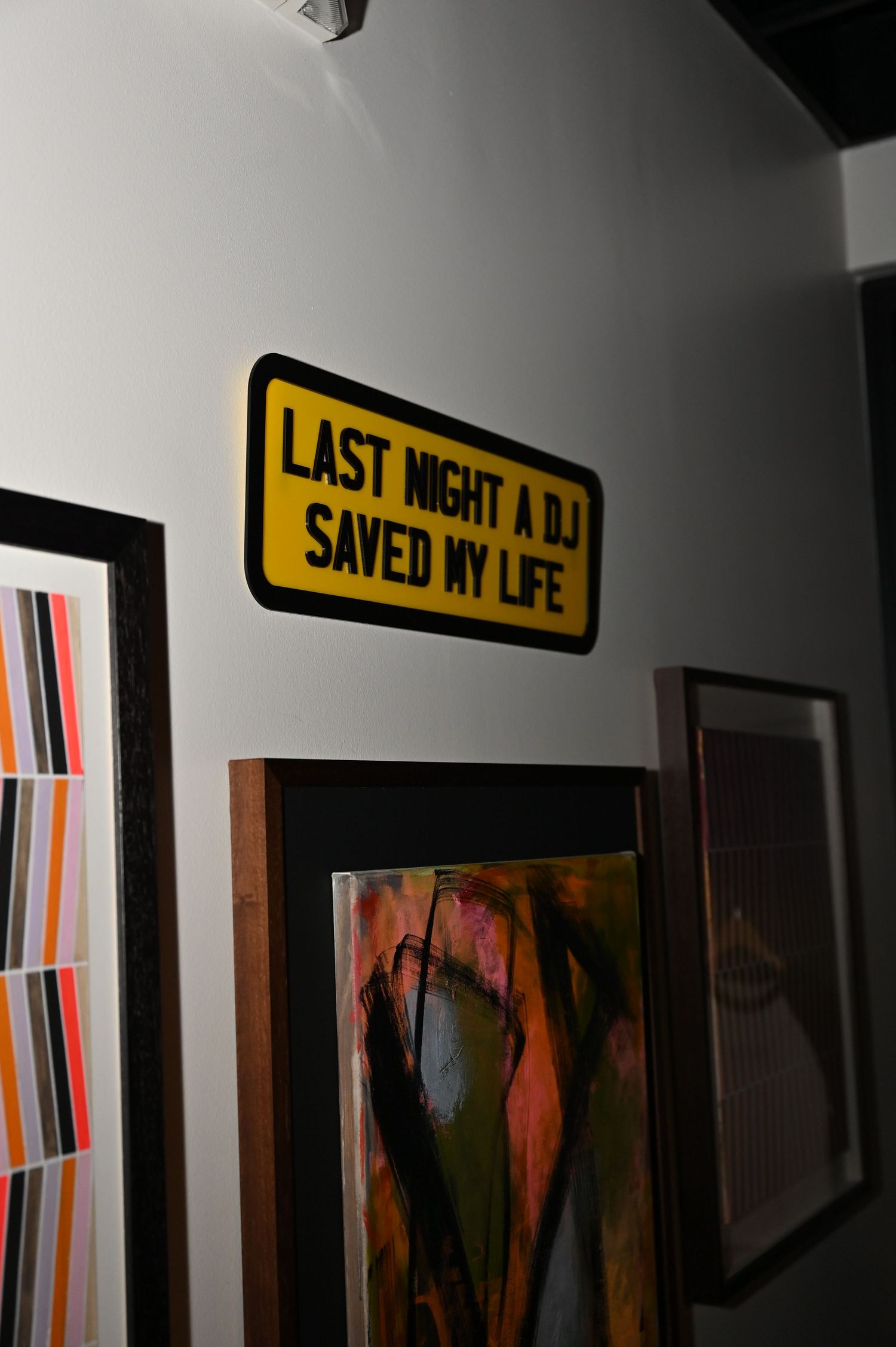 Yellow sign reading Last Night A DJ Saved My Life mounted on gallery wall surrounded by colorful framed artwork.