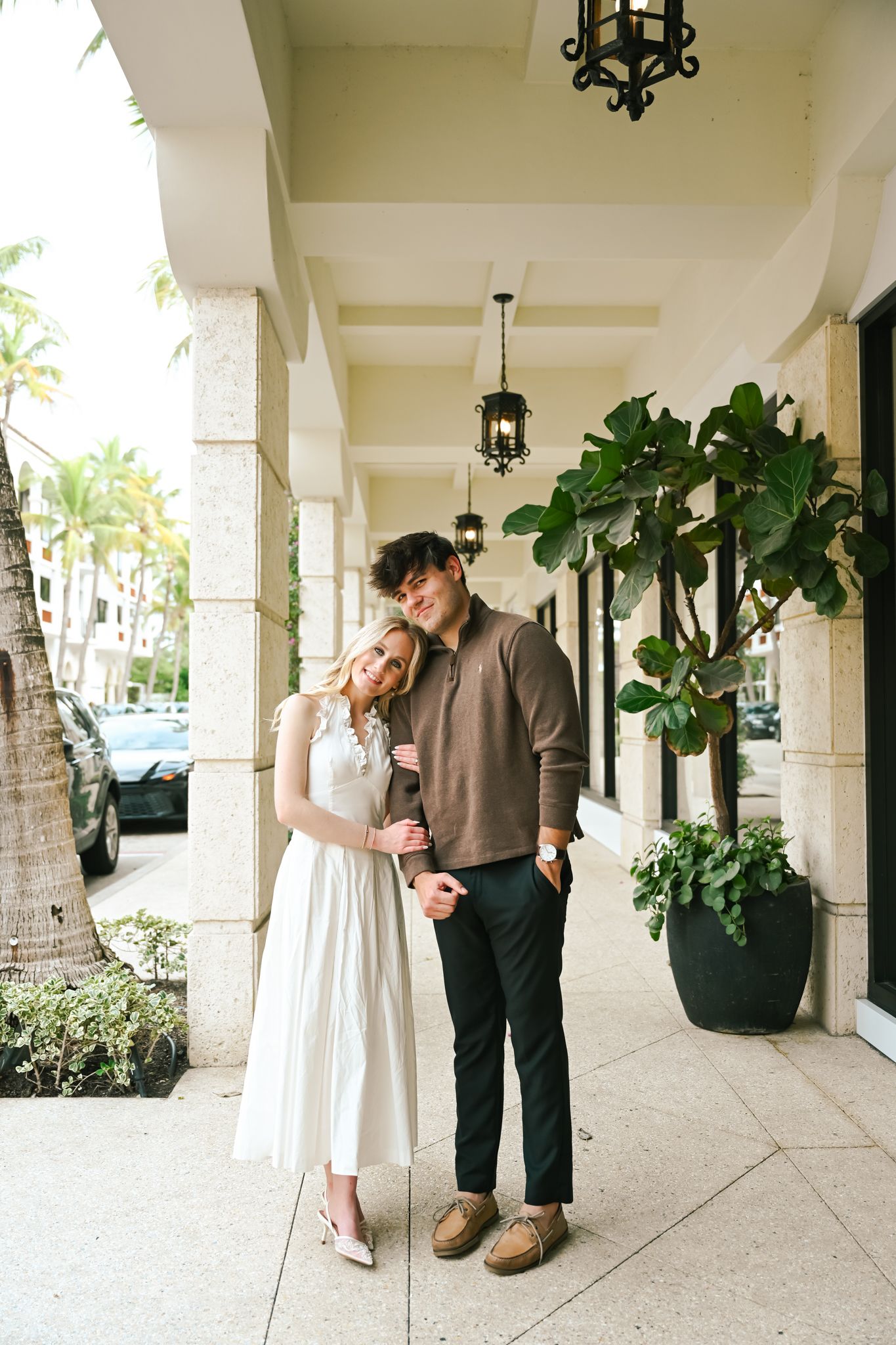 Couple posing under elegant covered walkway with hanging lanterns, woman in white dress, man in brown polo and black pants.
