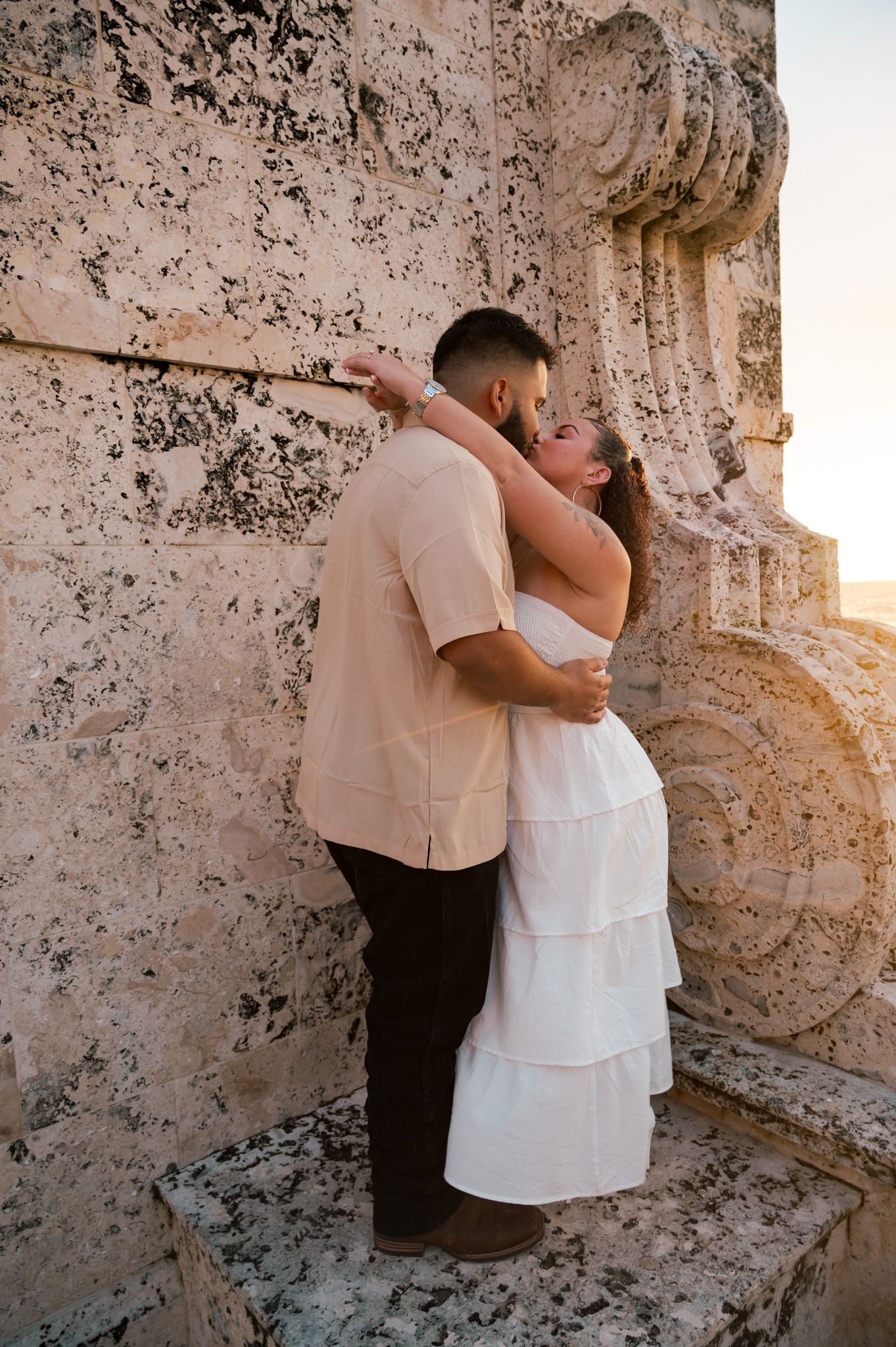 Couple sharing a romantic kiss against an ancient stone wall, woman in white tiered dress, man in beige shirt at sunset.