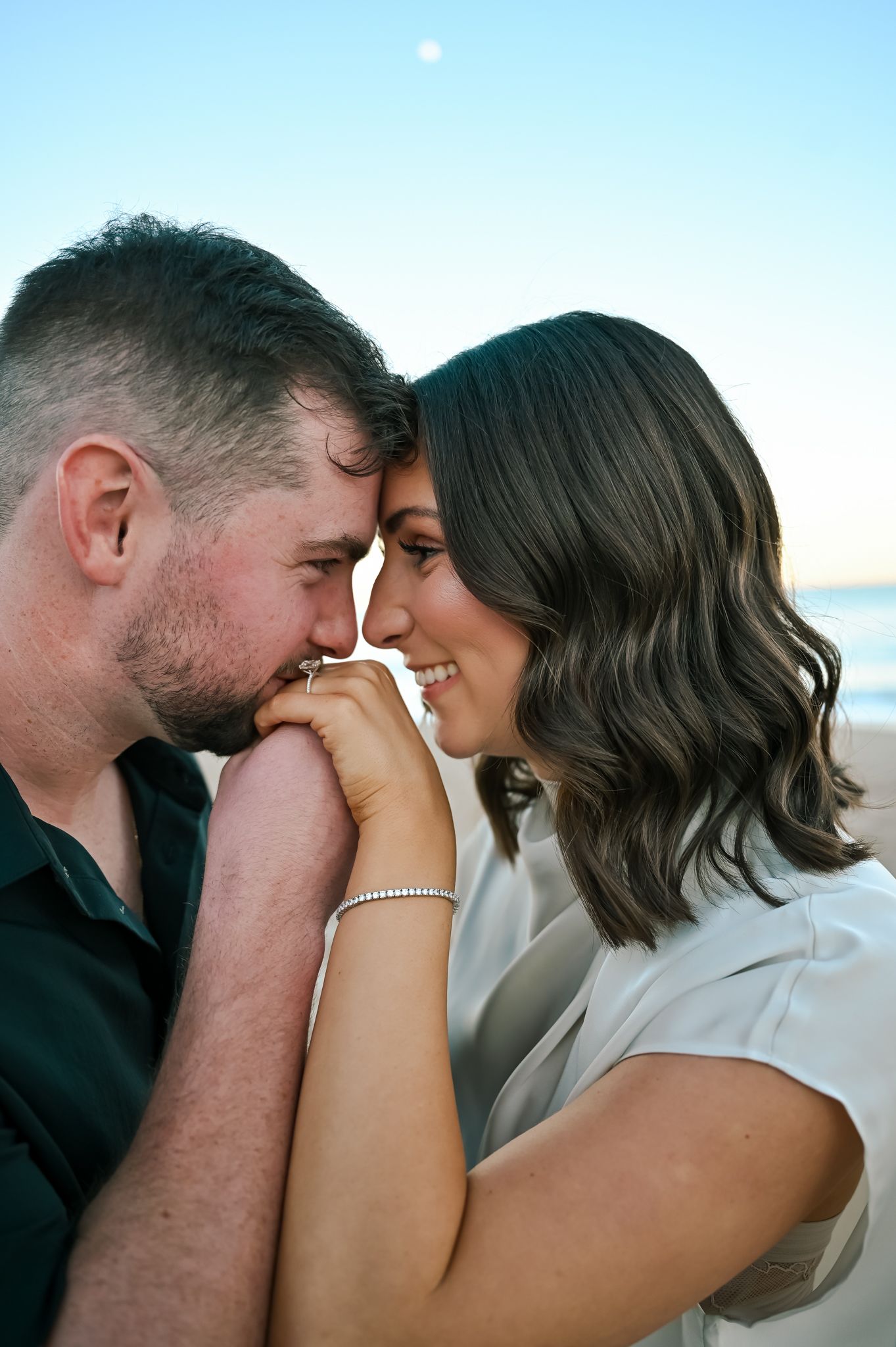 A couple shares a romantic moment on the beach, touching foreheads and smiling, the woman wearing a diamond ring.
