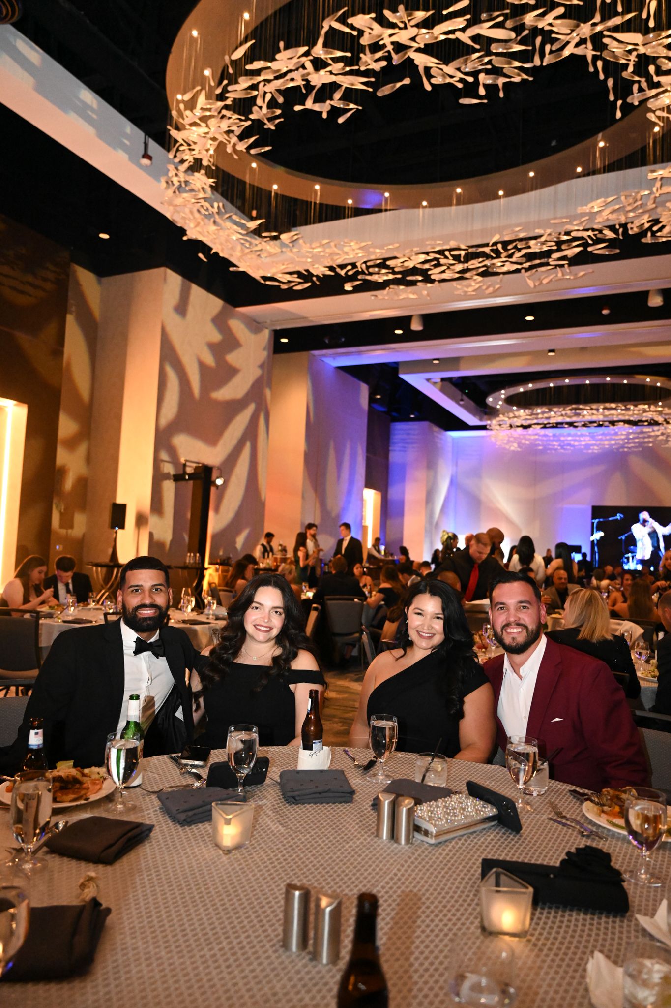 Four guests smile at a table during an elegant gala event with chandelier lighting in a grand ballroom.