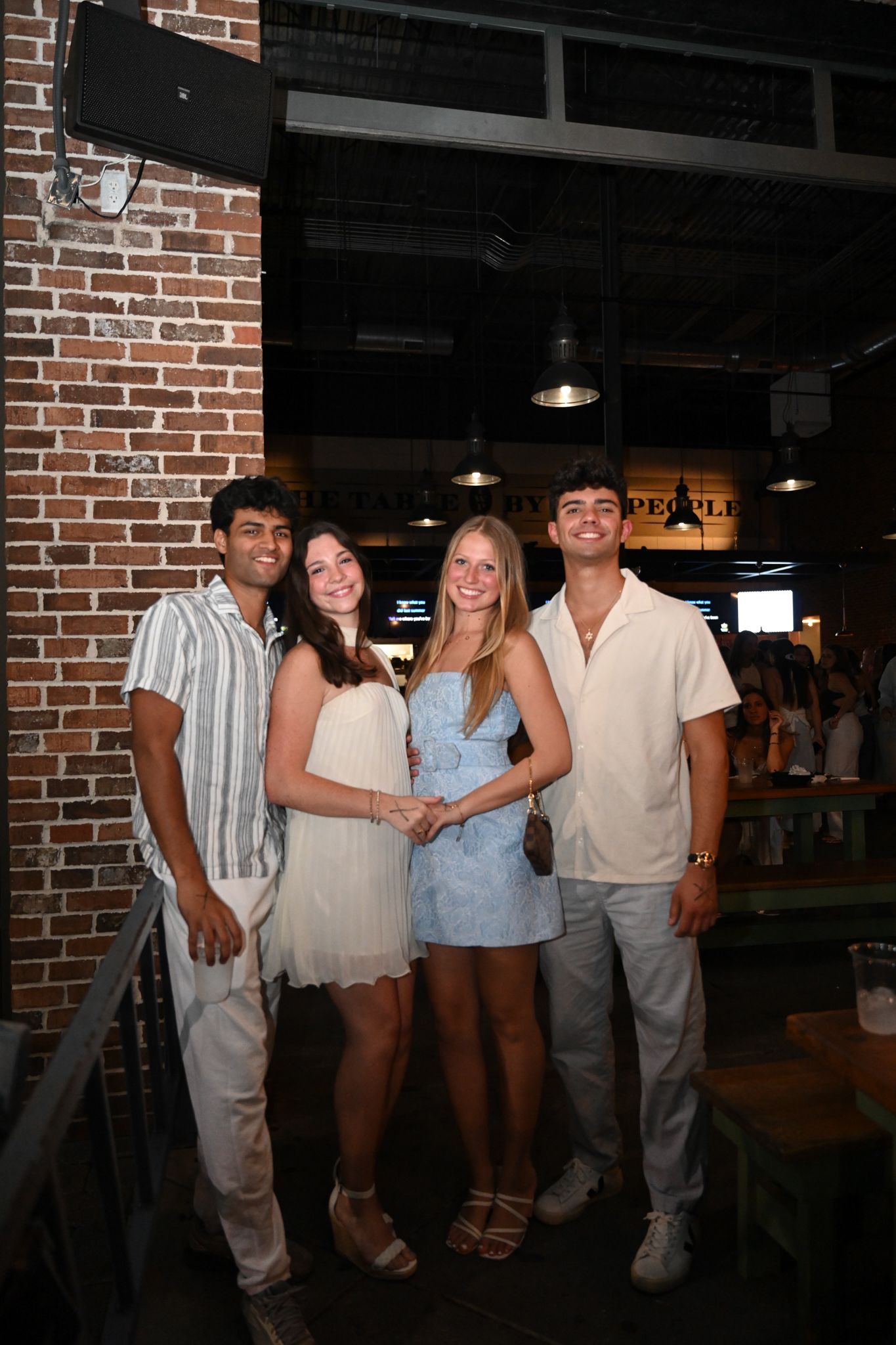 Four young friends smiling together at a casual indoor venue with exposed brick walls and pendant lights.