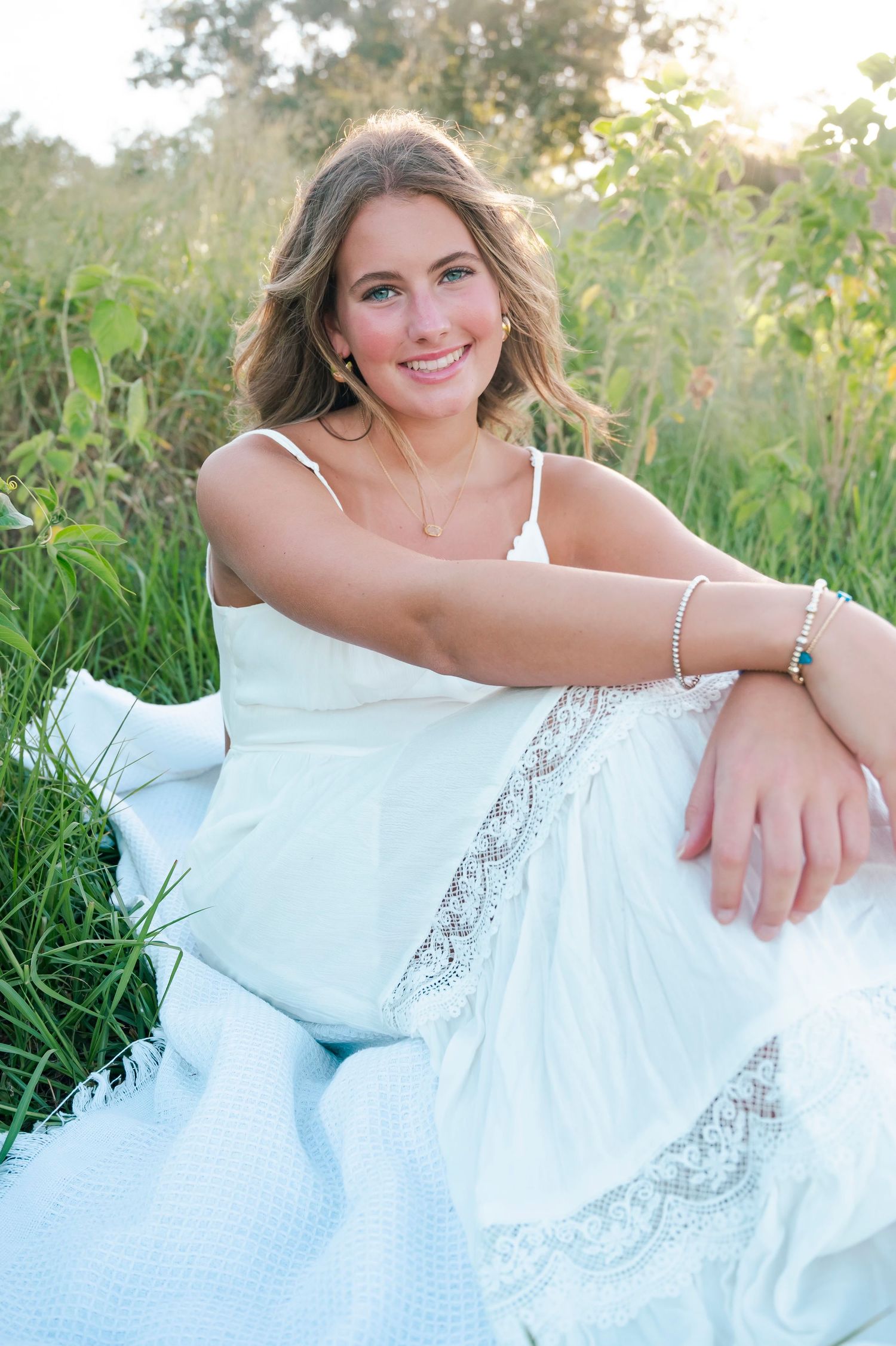 Young woman in white lace dress sitting in green field, smiling outdoors during golden hour.