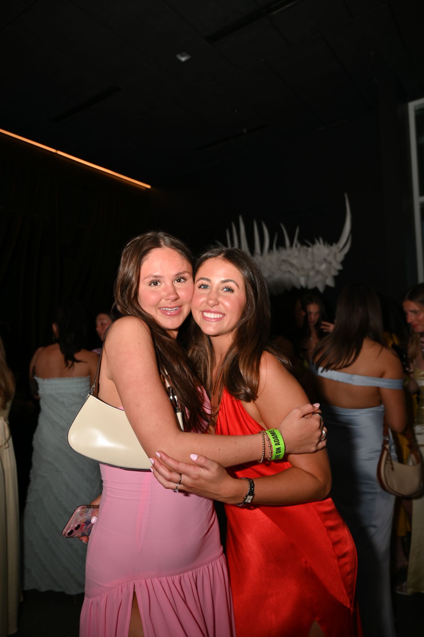 Two young women in elegant dresses share a warm hug at a formal event, smiling for the camera.