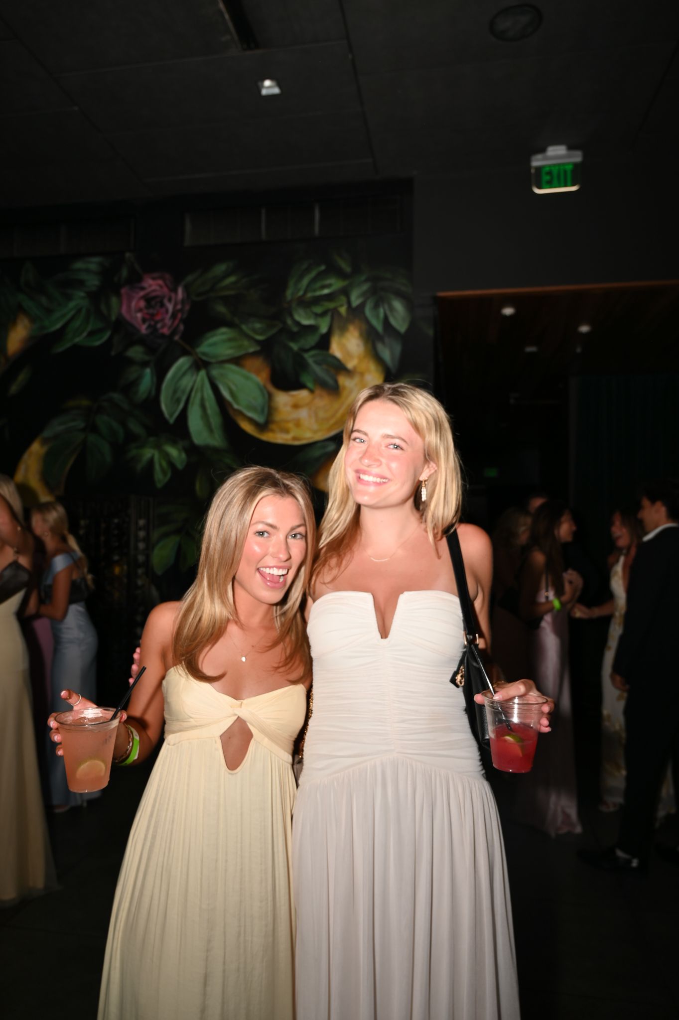 Two blonde women in elegant white and yellow gowns smile at a formal event, holding cocktails.