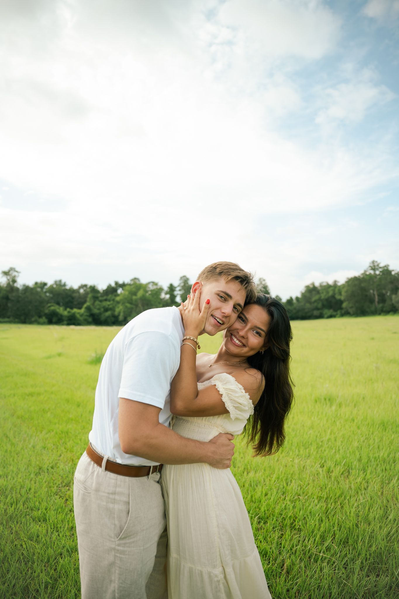 Happy couple embracing in a lush green field, wearing white outfits, smiling under a bright blue sky.