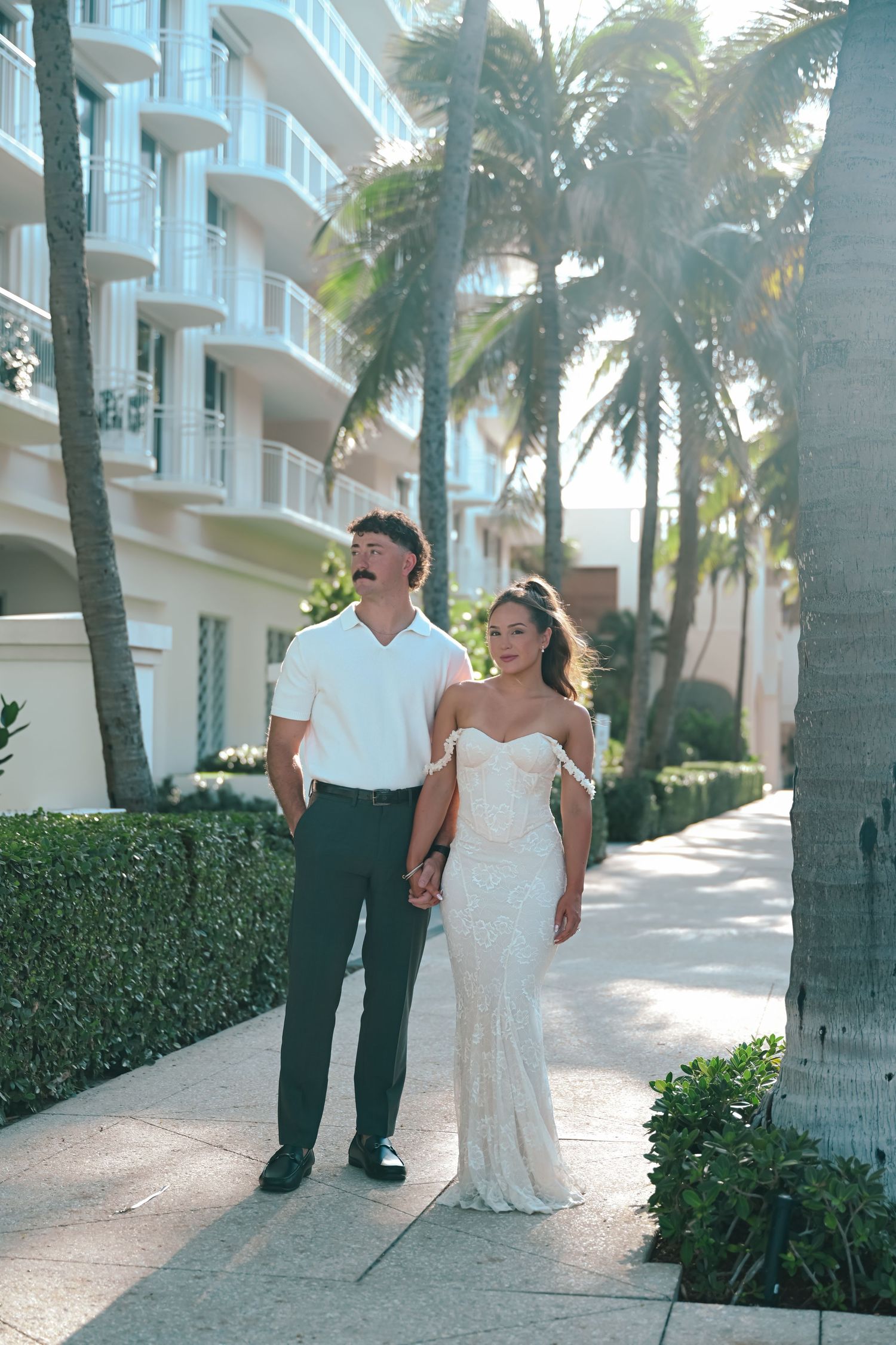 Couple in wedding attire walking hand-in-hand along a tropical resort pathway lined with palm trees and lush hedges.