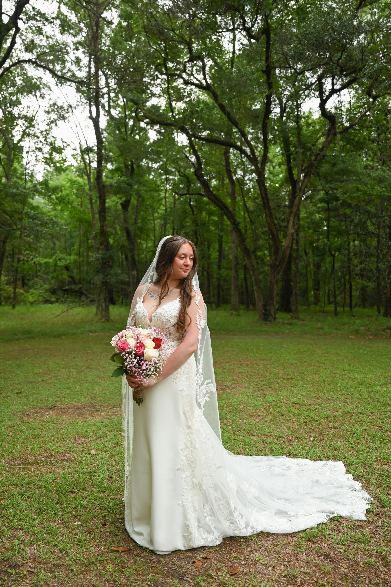 Bride in lace wedding gown with veil holds pink bouquet in lush green forest clearing.