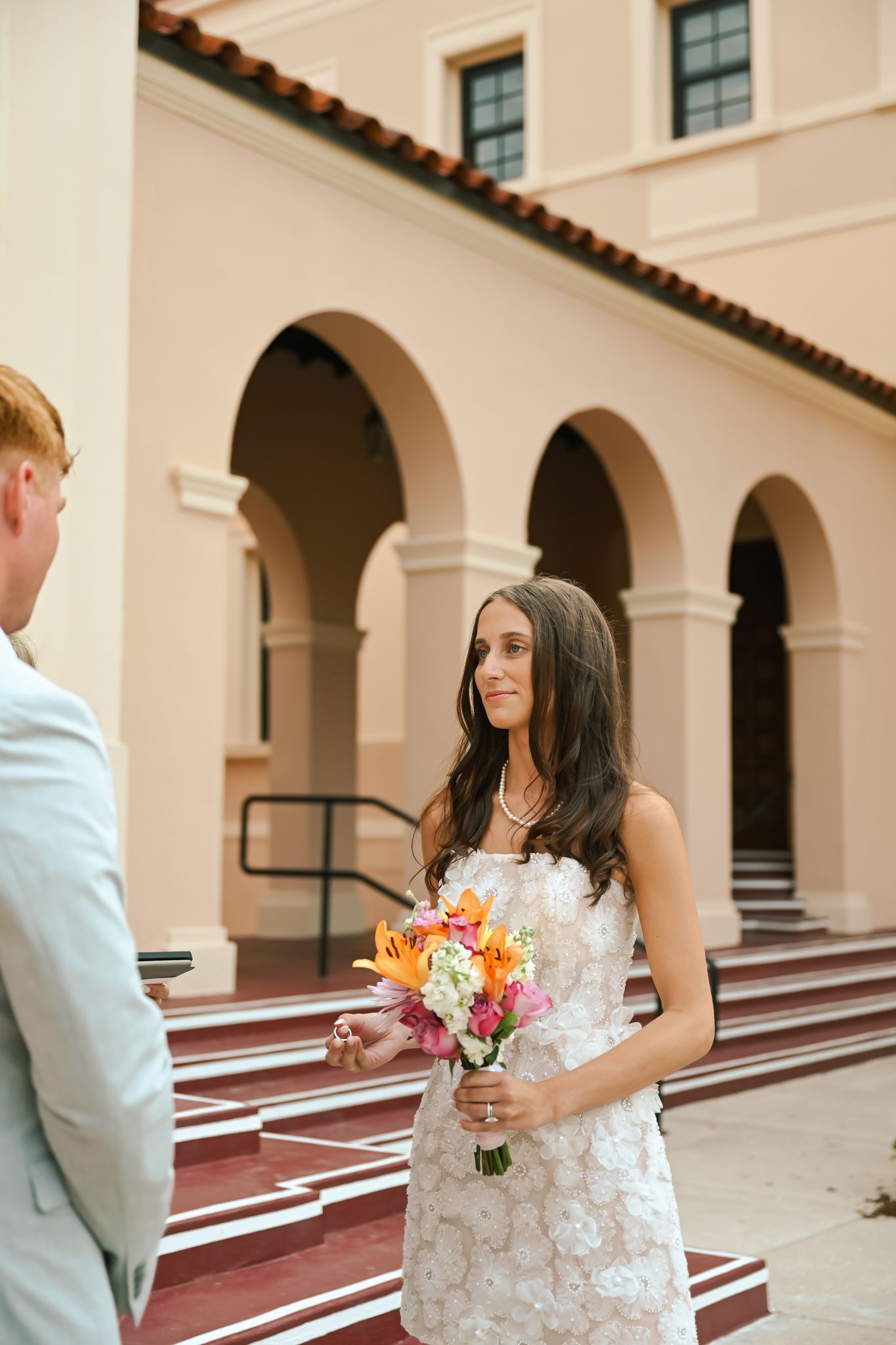 Bride in lace gown holds colorful bouquet during first look outside elegant Spanish-style building.
