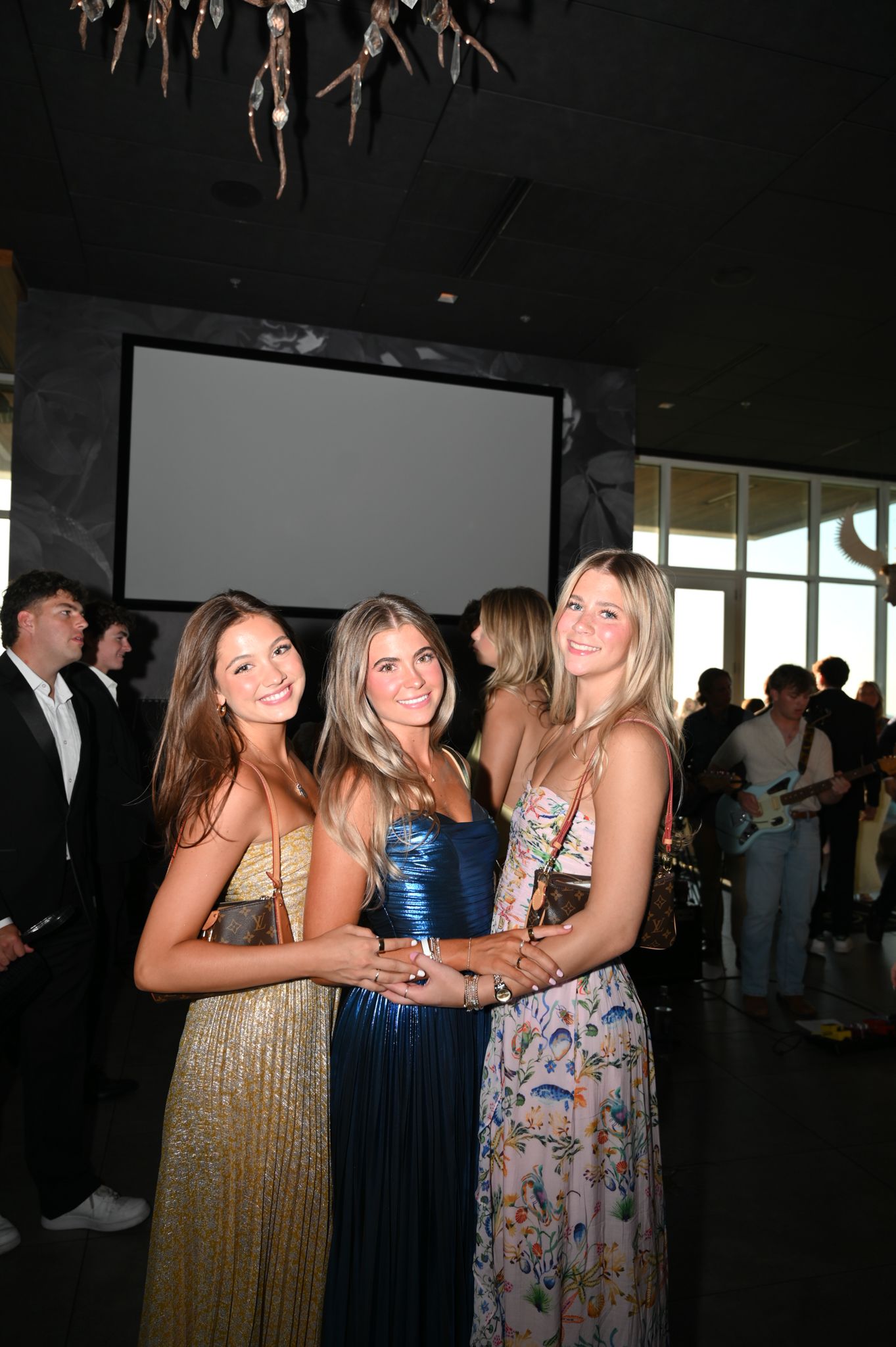 Three young women in elegant formal gowns smile together at an indoor event with guests and a projection screen.
