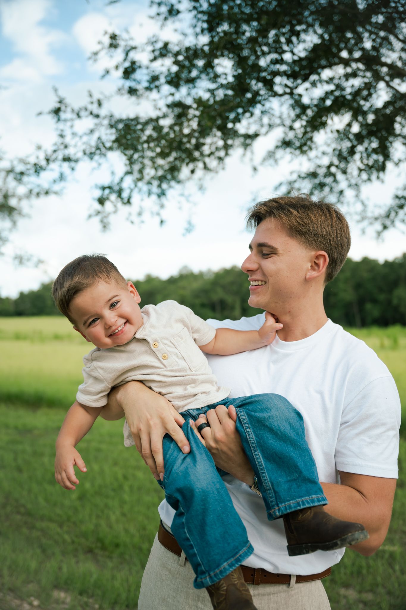 Young father in white t-shirt laughing while holding his smiling toddler son outdoors in a green field.