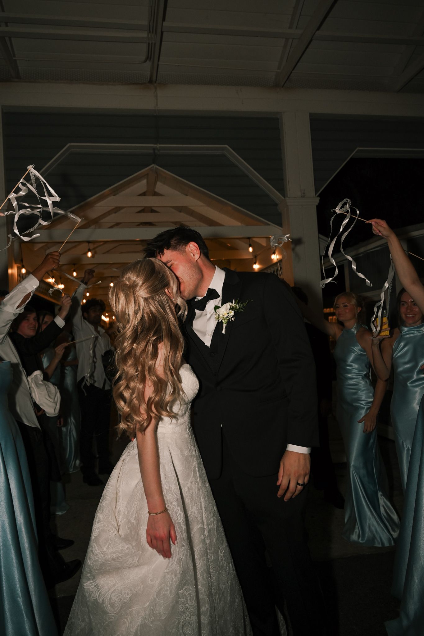 Bride and groom share a kiss during wedding reception exit with guests waving ribbon wands in elegant venue.