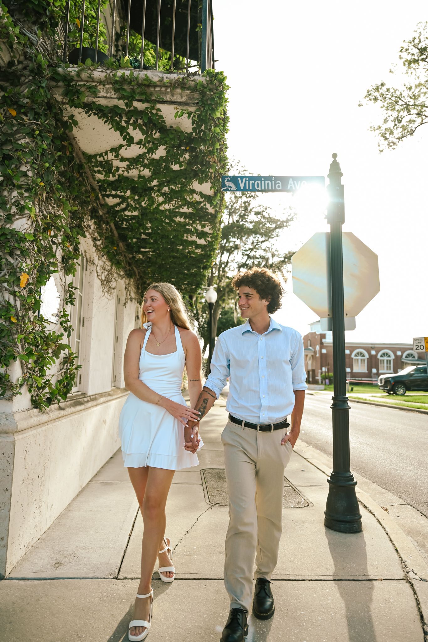 A smiling couple holds hands while walking on a sunny sidewalk near a Virginia Ave street sign, surrounded by ivy-covered buildings.