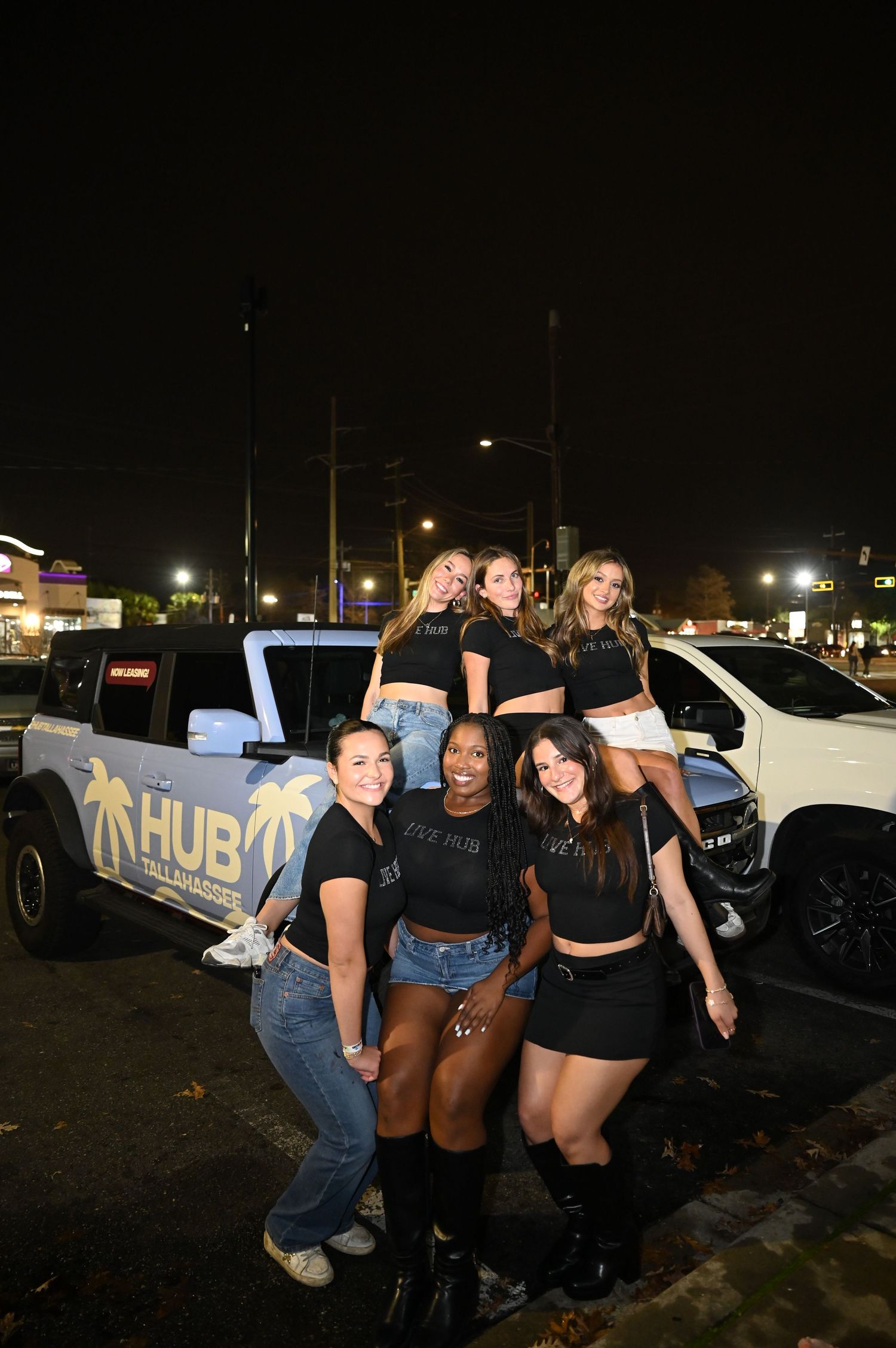 Six young women posing together at night in front of a white HUB-branded Jeep in a parking lot.