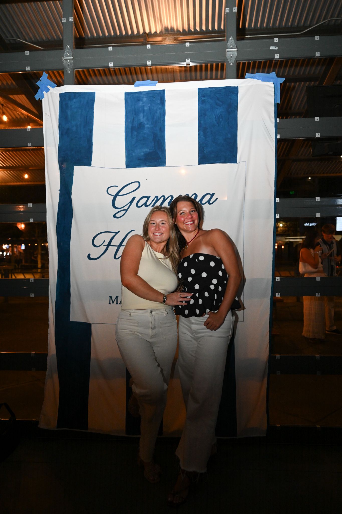 Two women pose together in front of a blue and white Gamma striped banner at an indoor event.