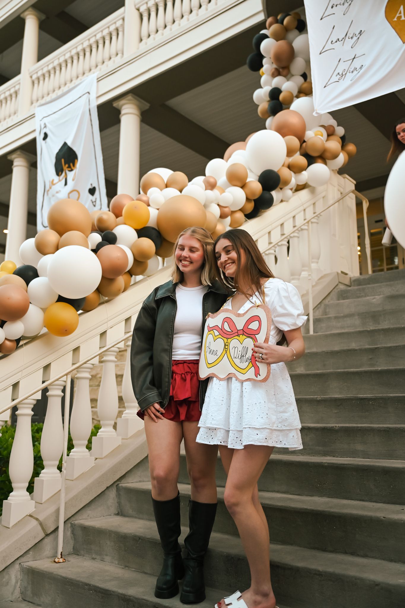 Two smiling young women pose on decorated stairs with graduation balloons at a celebratory event.