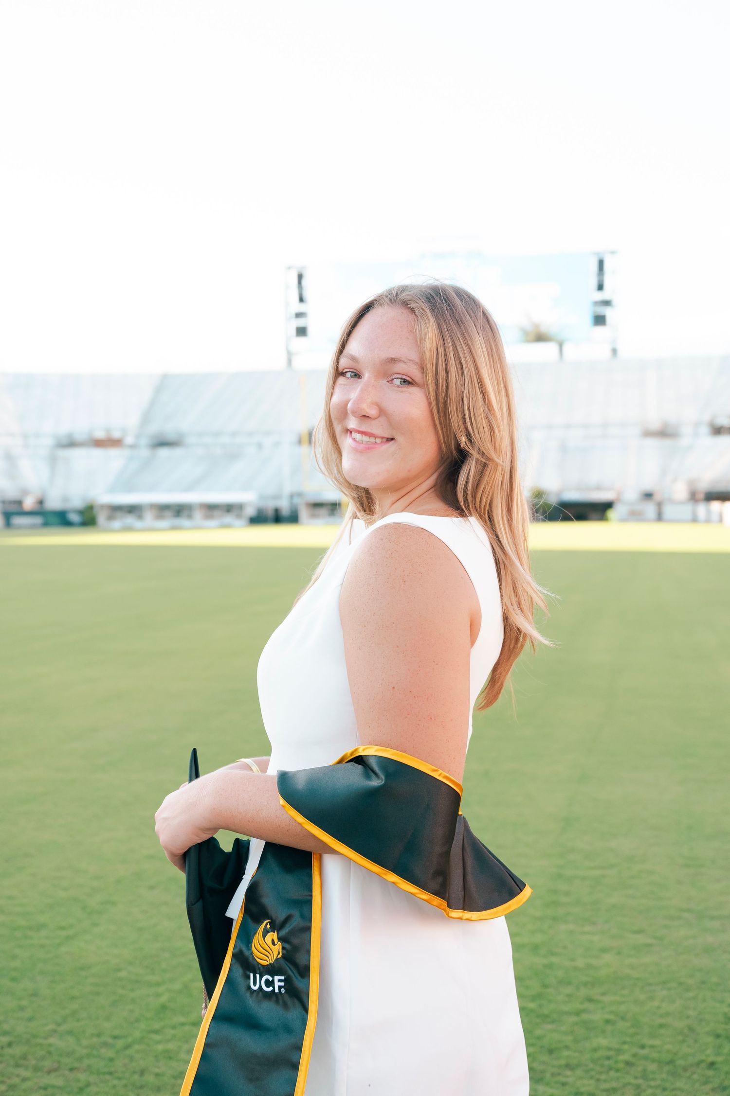 Graduate in white dress poses with UCF graduation stole on sports field.