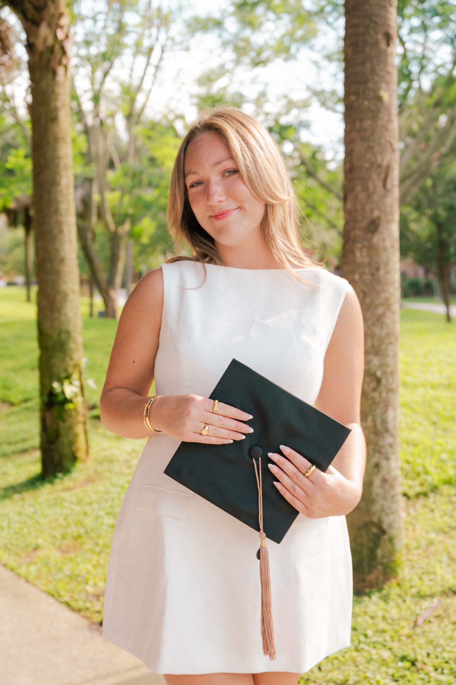 Graduate in white dress holds diploma and tassel while standing in wooded outdoor setting.