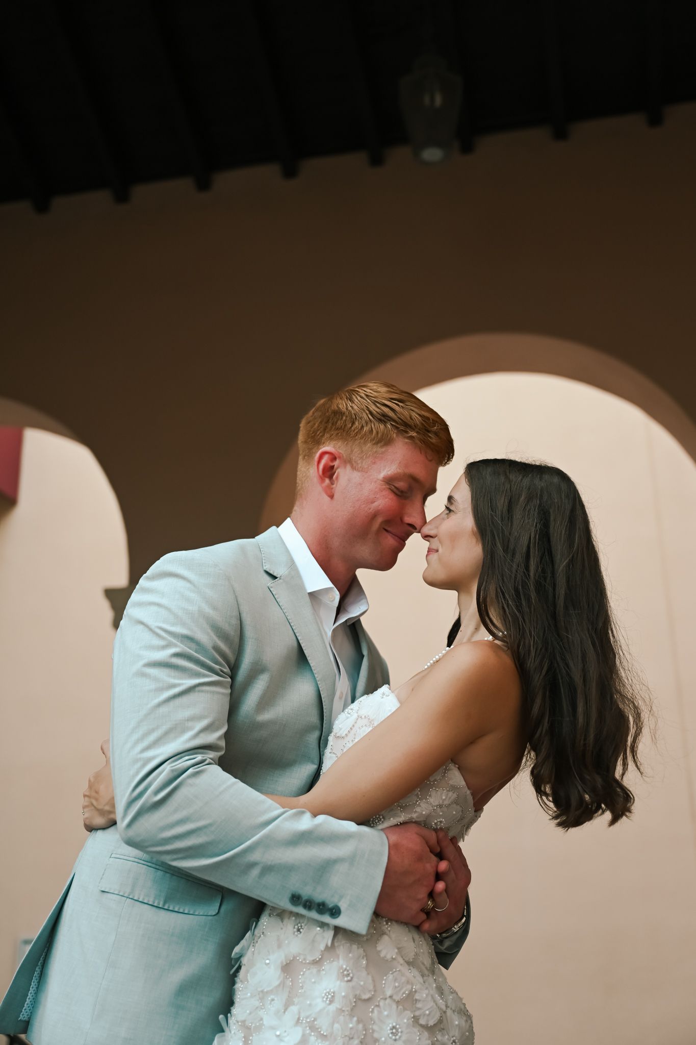 A bride and groom share a romantic first dance moment, nearly kissing, as the groom wears a light blue suit.