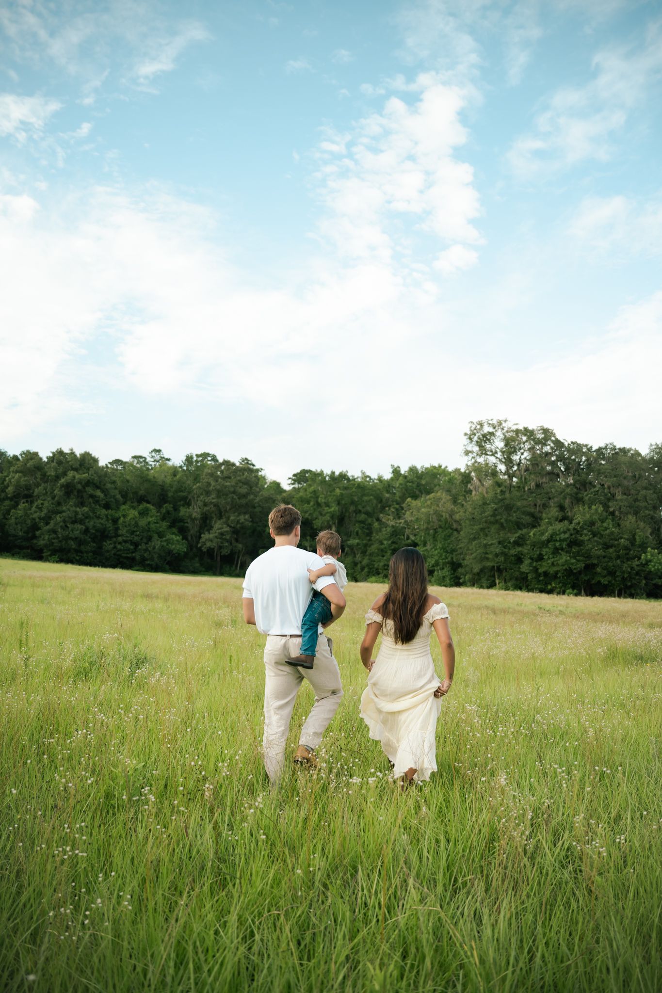 Family of three walking through lush green meadow, parents carrying child, trees and blue sky in background.