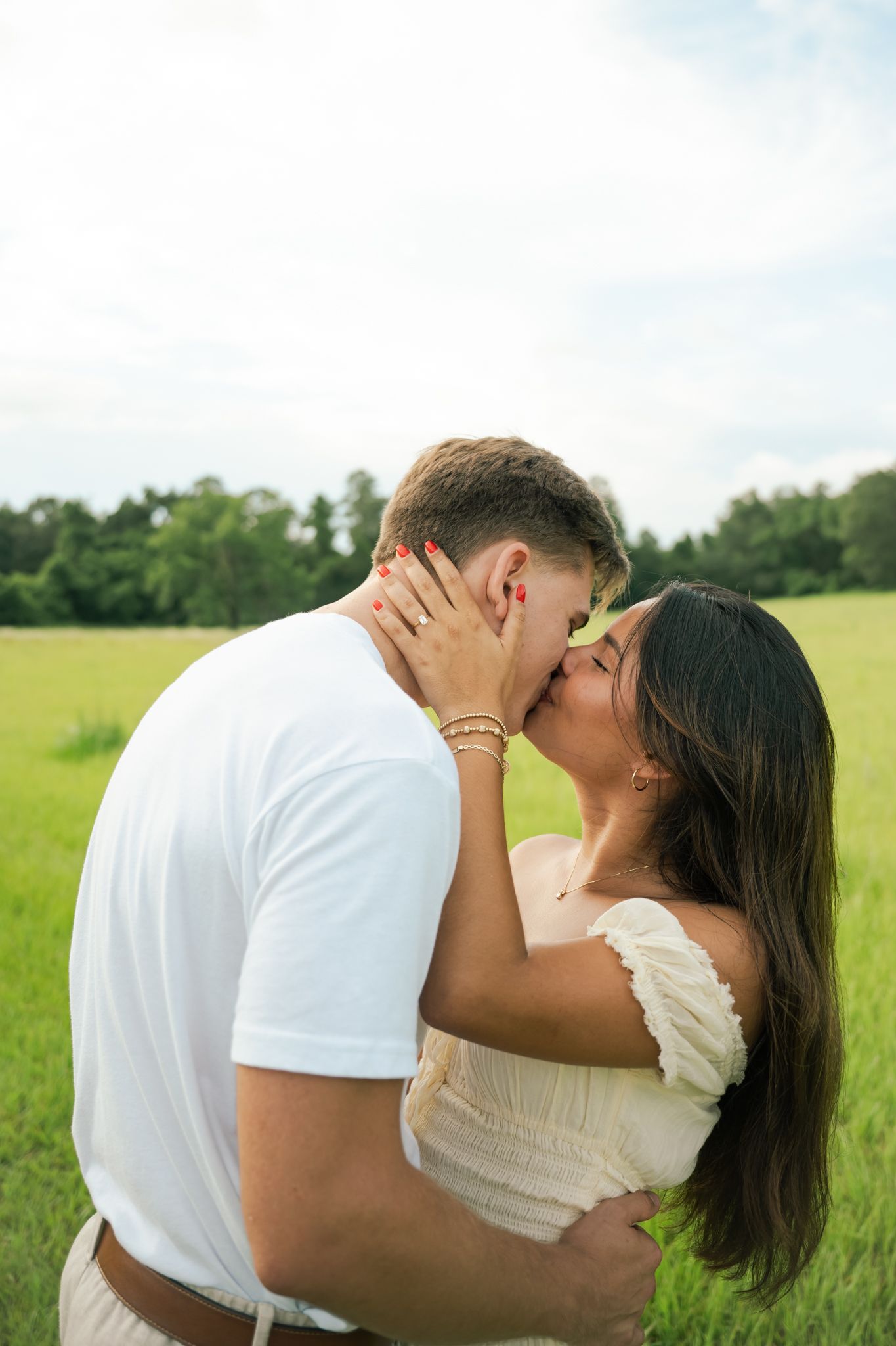 Couple sharing a romantic kiss in a lush green field, woman in white dress with red nails, man in white t-shirt.