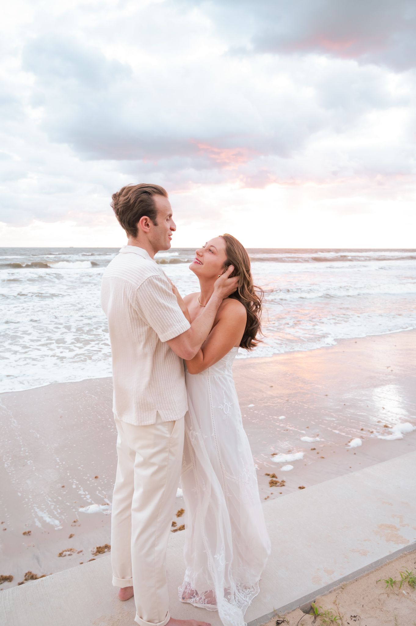 A couple embraces romantically on a beach at sunset, wearing white outfits with ocean waves in the background.