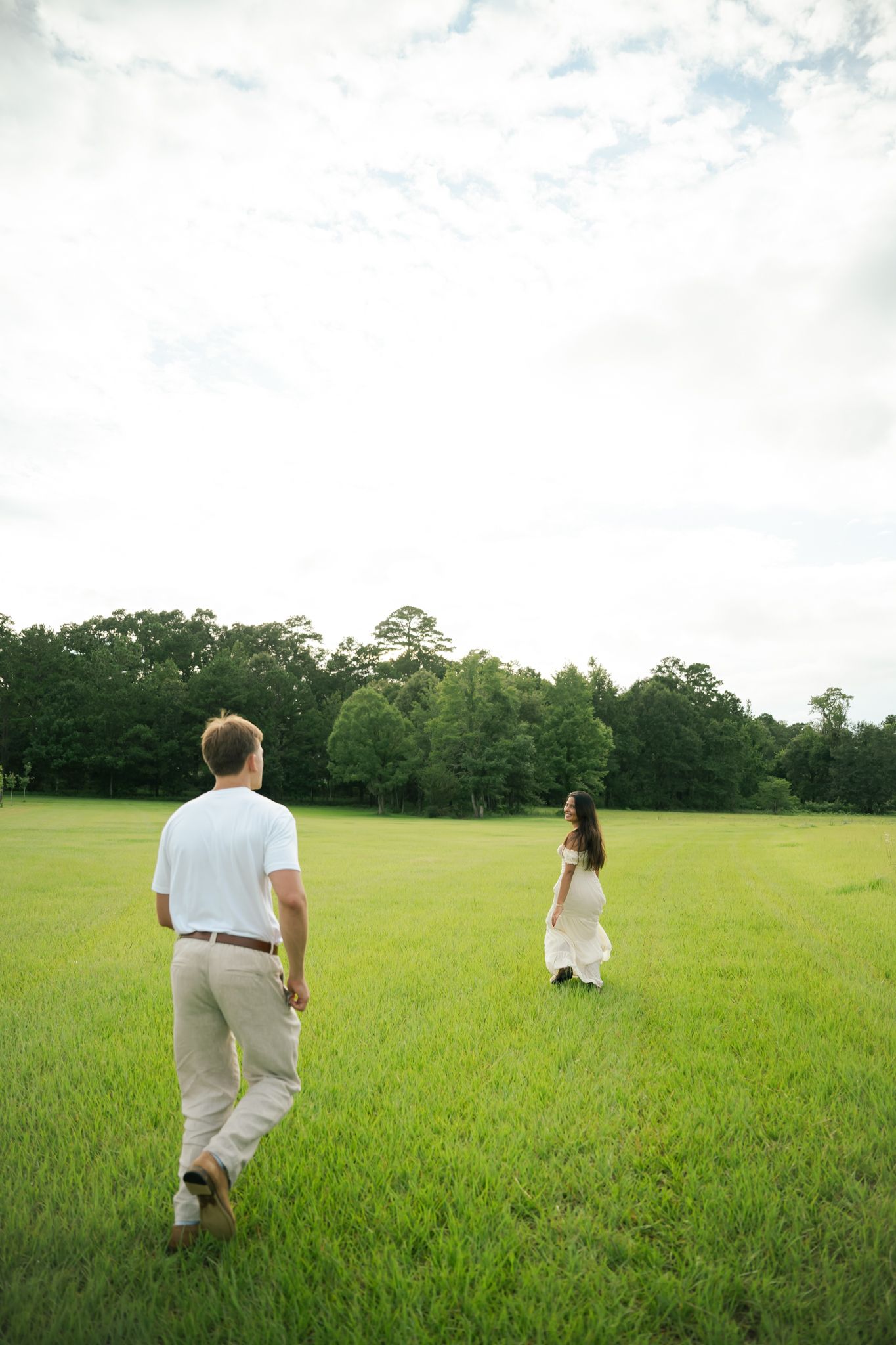 Couple in white attire walking through a lush green field with trees in the background.