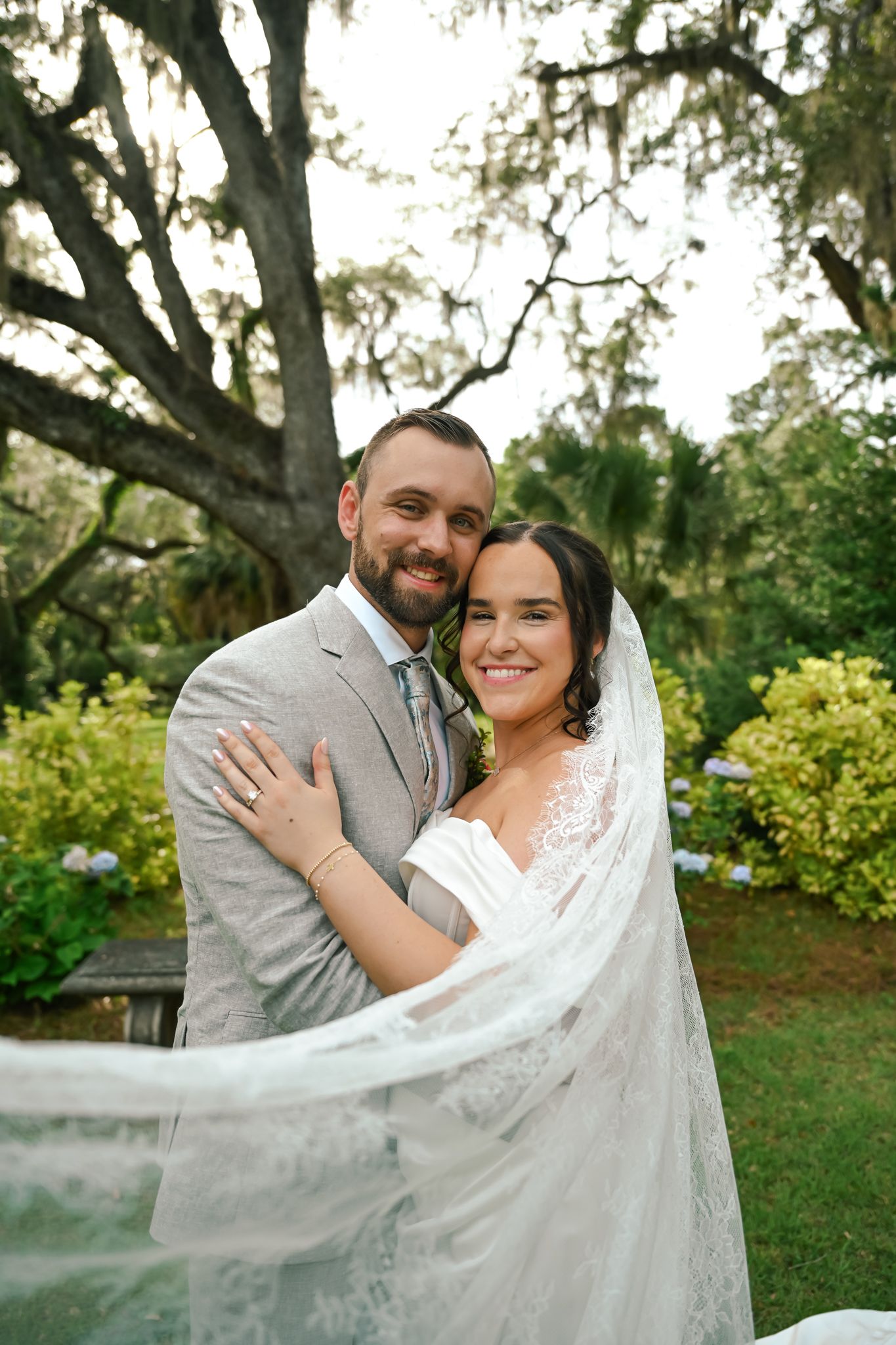 A smiling bride and groom embrace outdoors under a large oak tree, with the bride's veil flowing in the breeze.