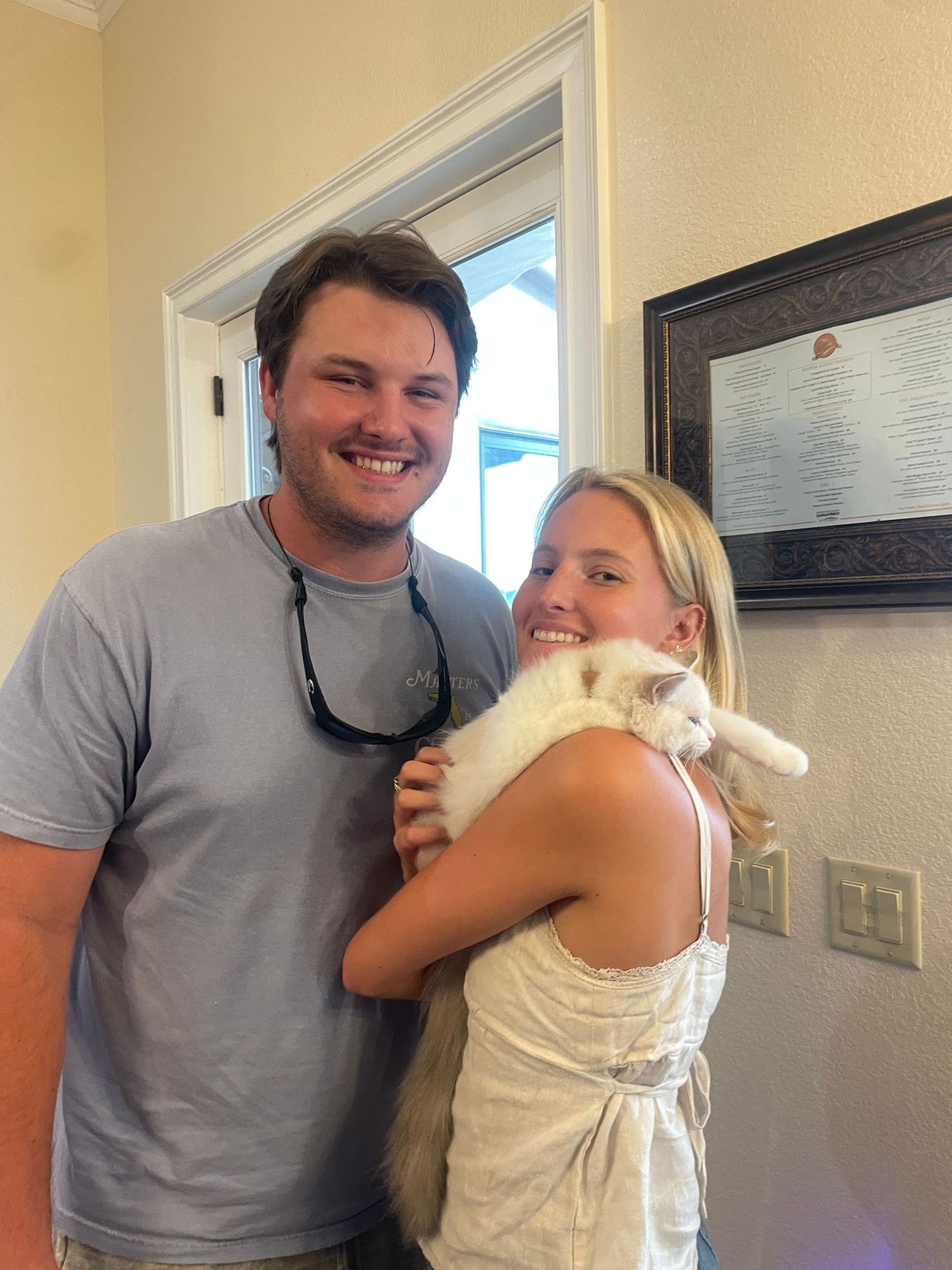 Smiling couple holding a fluffy white cat indoors, posing together in a bright room with a framed document on the wall.