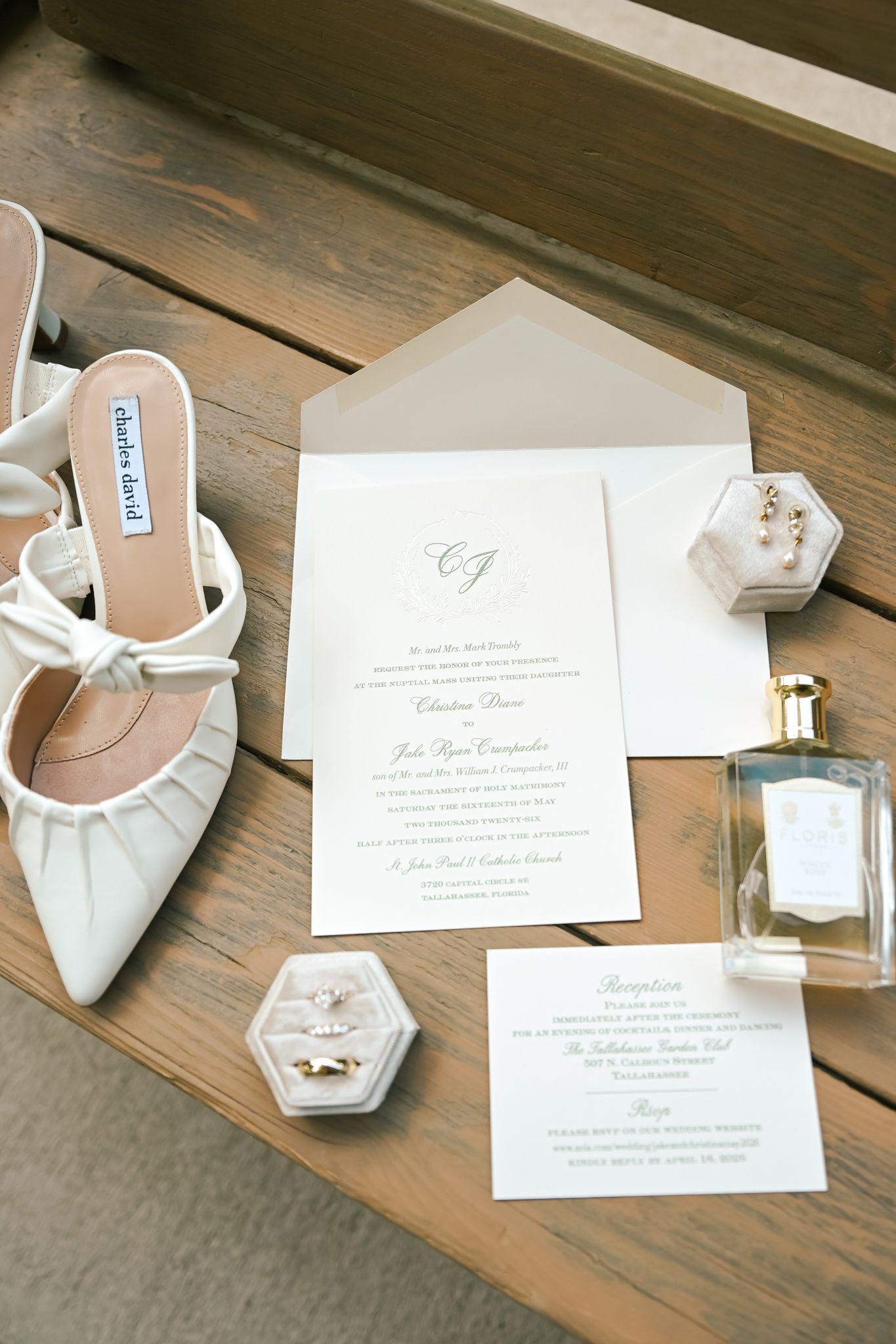 Elegant wedding flat lay with white bow heels, invitation suite, velvet ring box, and perfume bottle on wooden surface.