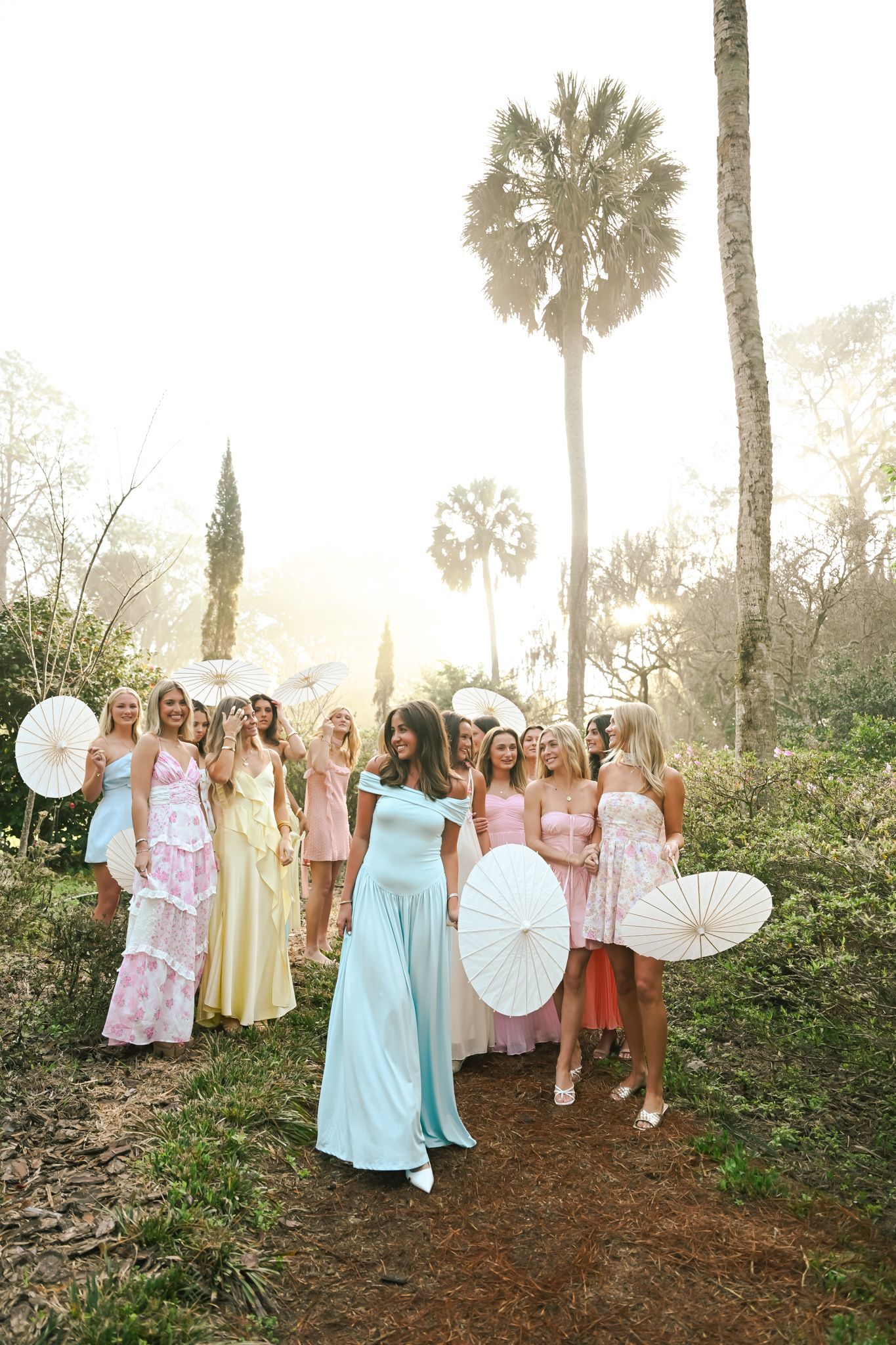 Bride in blue gown leads bridesmaids in pastel dresses holding white parasols outdoors near palm trees in golden sunlight.