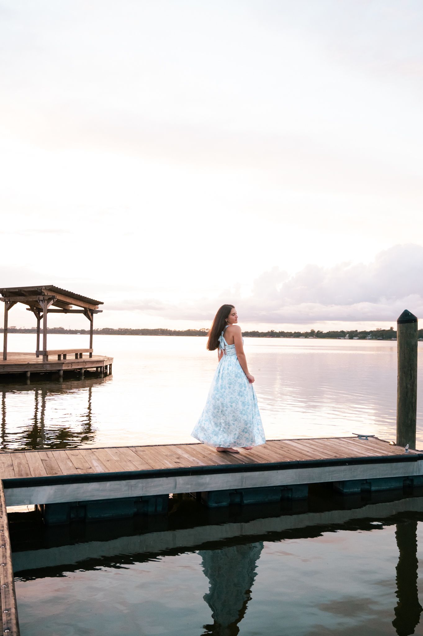 Woman in floral maxi dress standing on wooden dock at sunset over calm lake with covered pier in background.