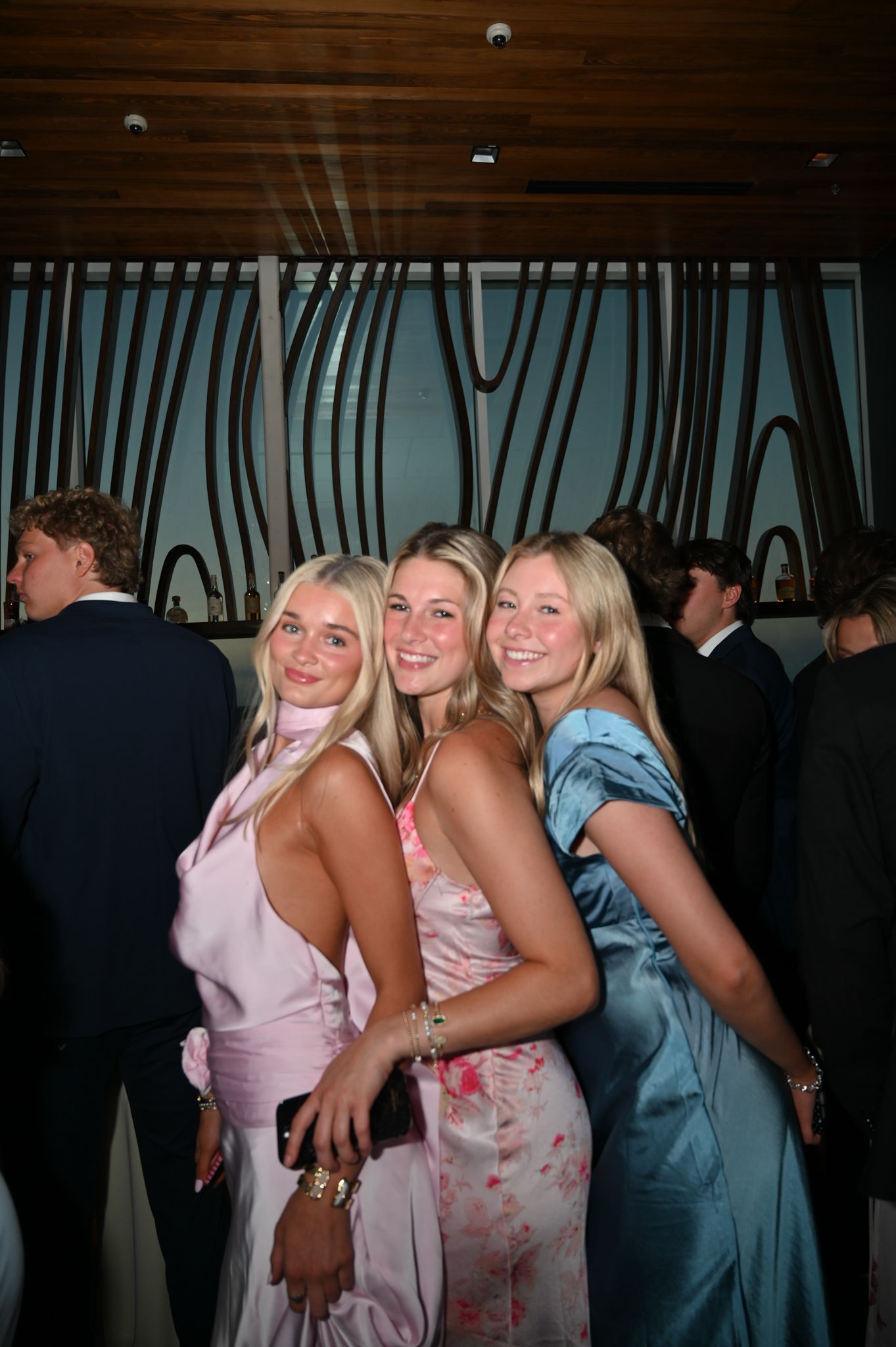 Three young women in elegant formal gowns pose together at a prom or formal event.