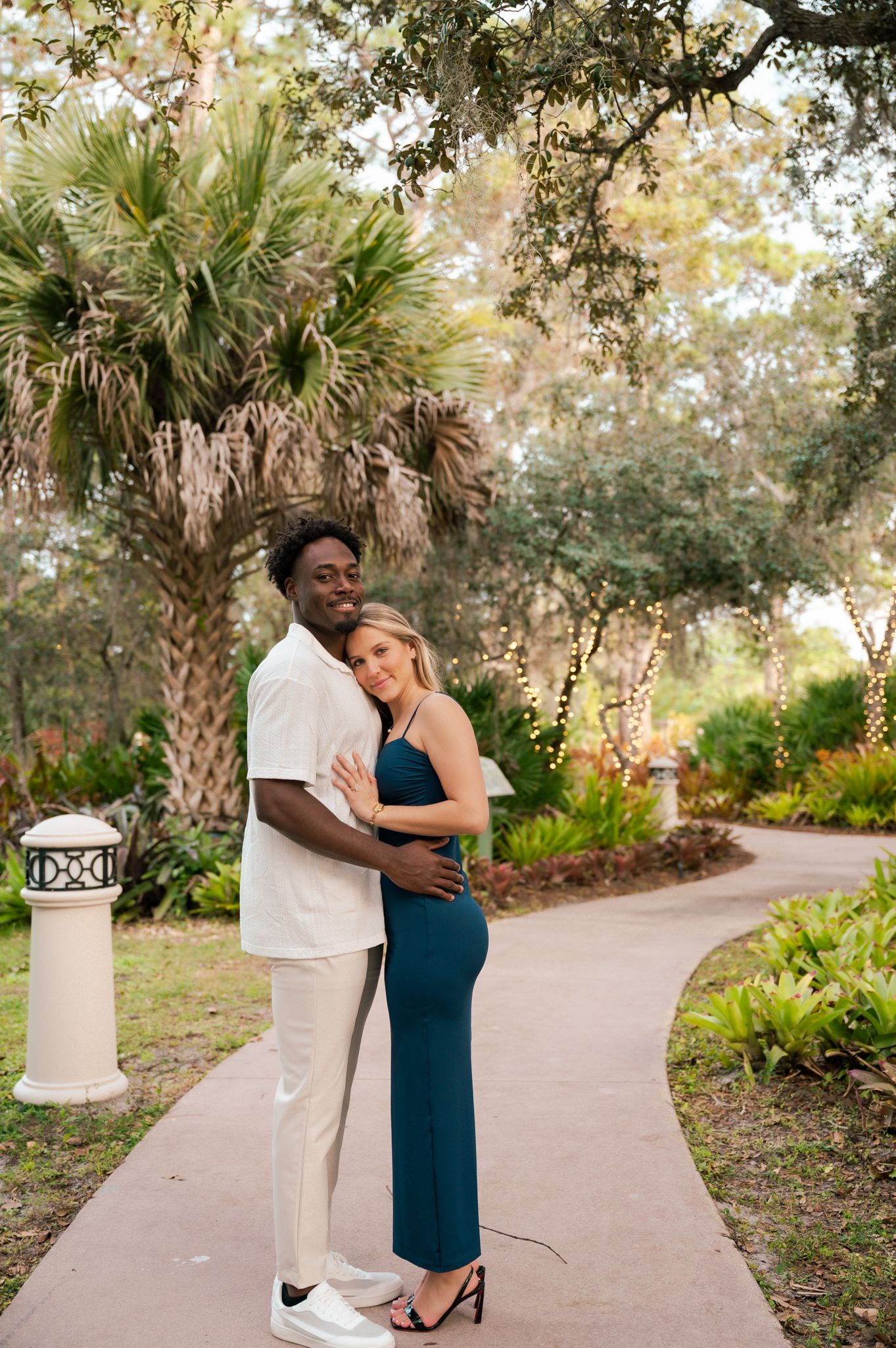A couple embraces on a garden path, woman in teal dress, man in white outfit, surrounded by lush greenery and fairy lights.
