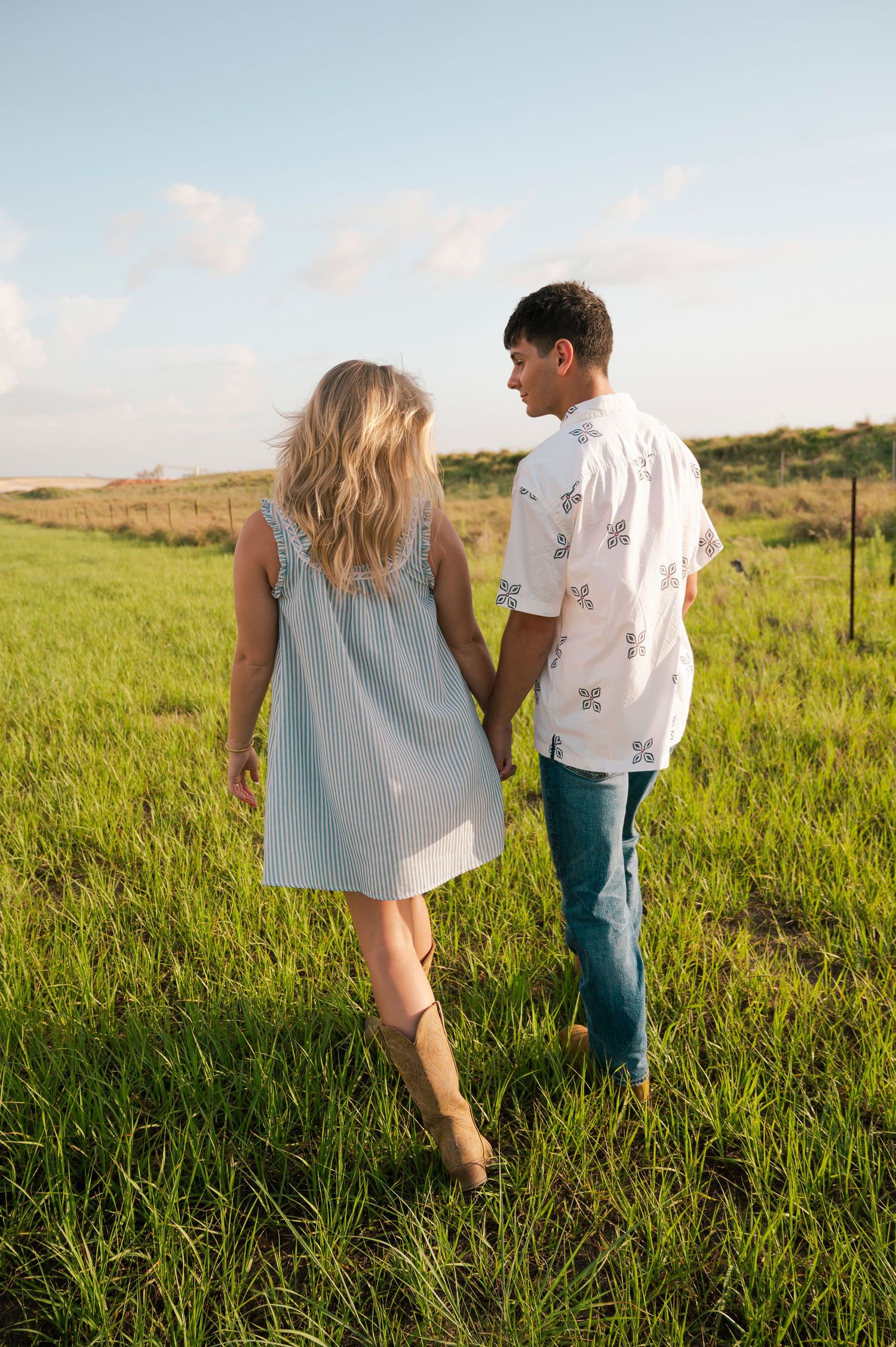 Couple holding hands walking through lush green field, woman in light blue dress and man in floral shirt and jeans.