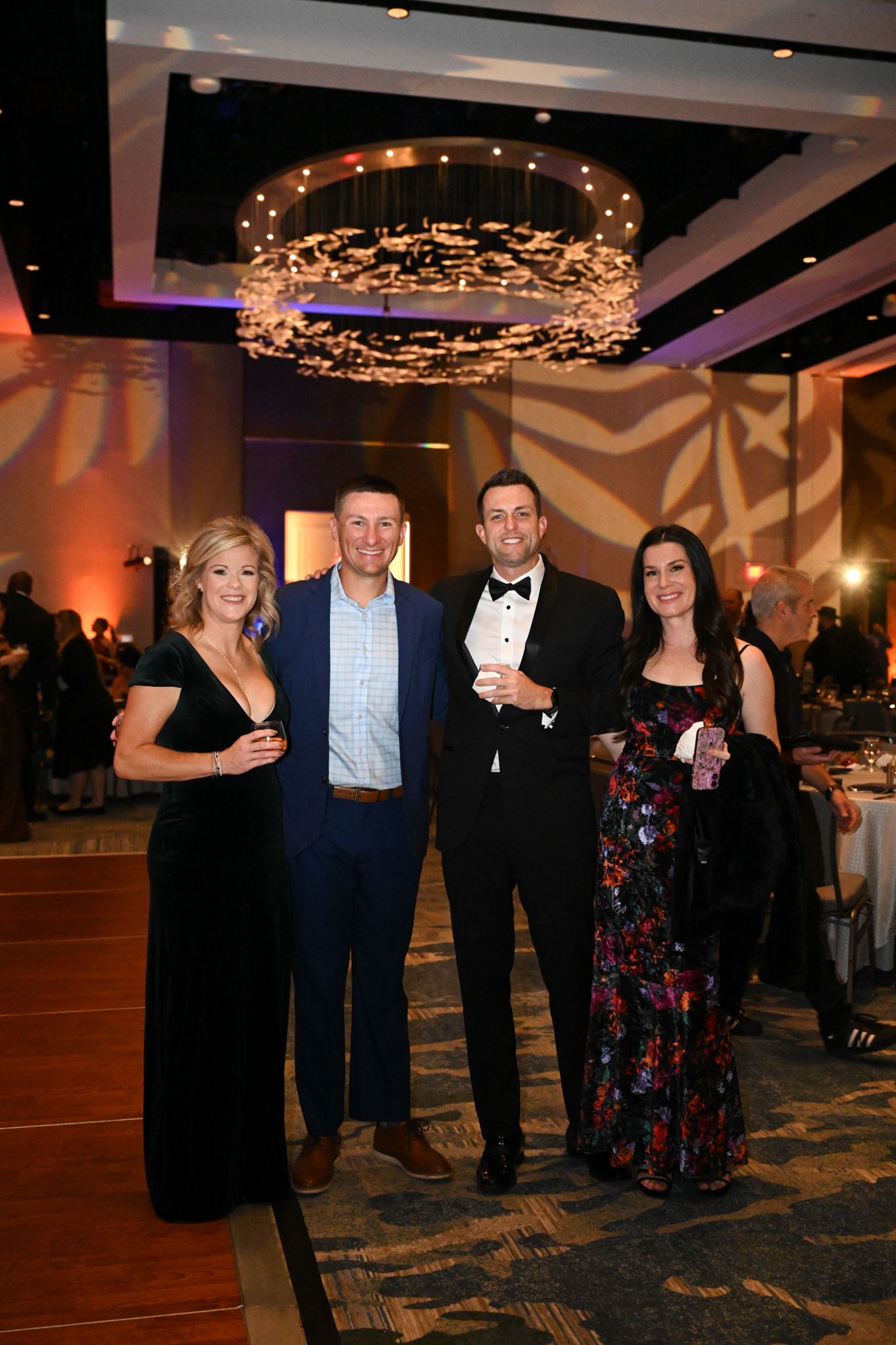 Four elegantly dressed guests posing together at a formal gala event in a grand ballroom with chandelier lighting.