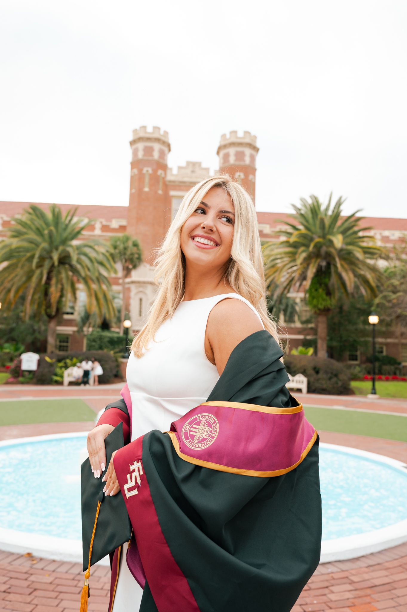 Graduate in white dress holding up academic regalia against a brick university building with palm trees.