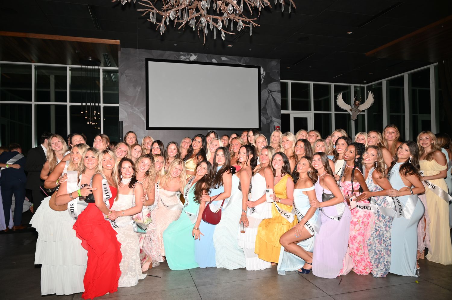Large group of women in colorful formal gowns posing together at an elegant indoor event venue.