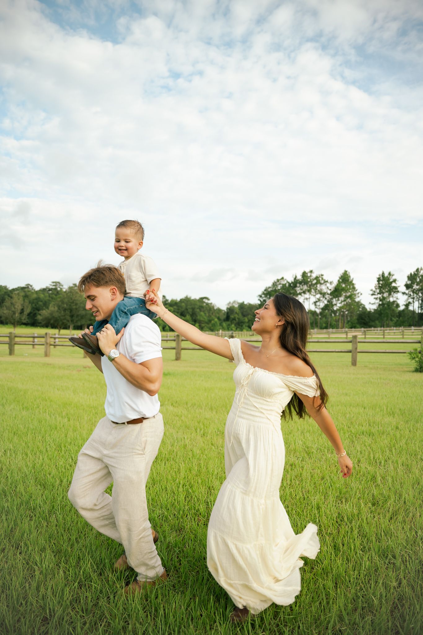 Happy family playing outdoors in green field, father lifting toddler on shoulders while mother in white dress reaches up smiling.