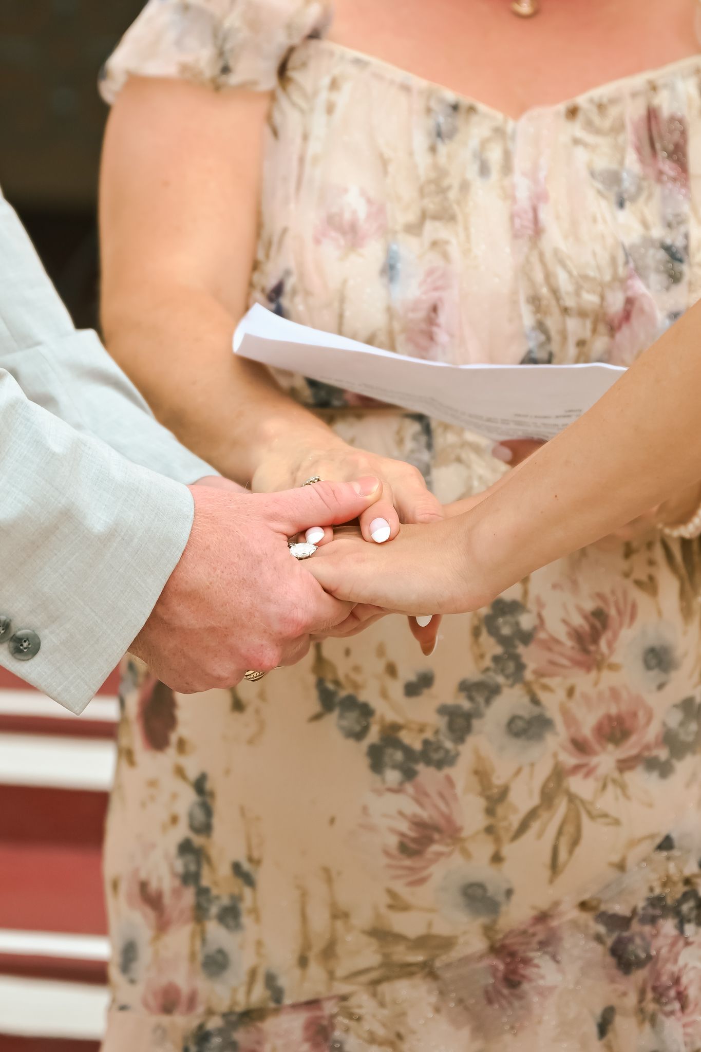 Couple exchanging wedding rings during outdoor ceremony, bride in floral dress holding hands with groom in light blue suit.