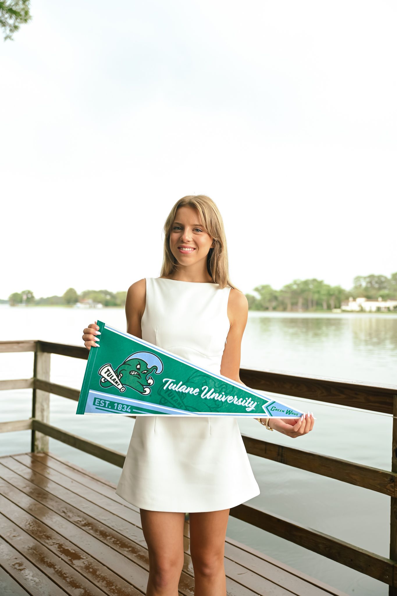 Young woman in white dress holding a Tulane University pennant on a wooden dock by the water.