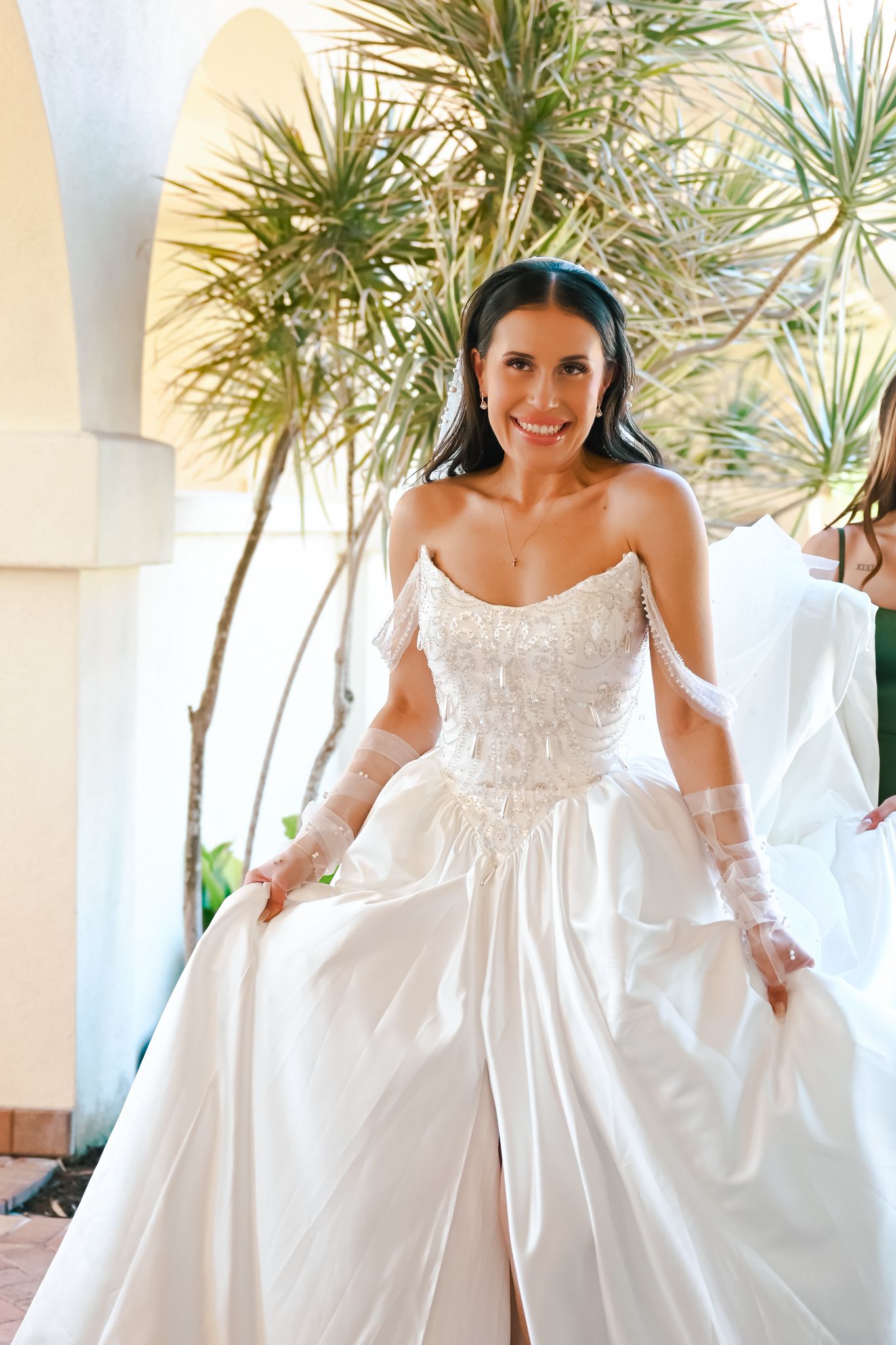 Smiling bride in elegant strapless white ball gown with lace gloves poses outdoors near tropical palms.