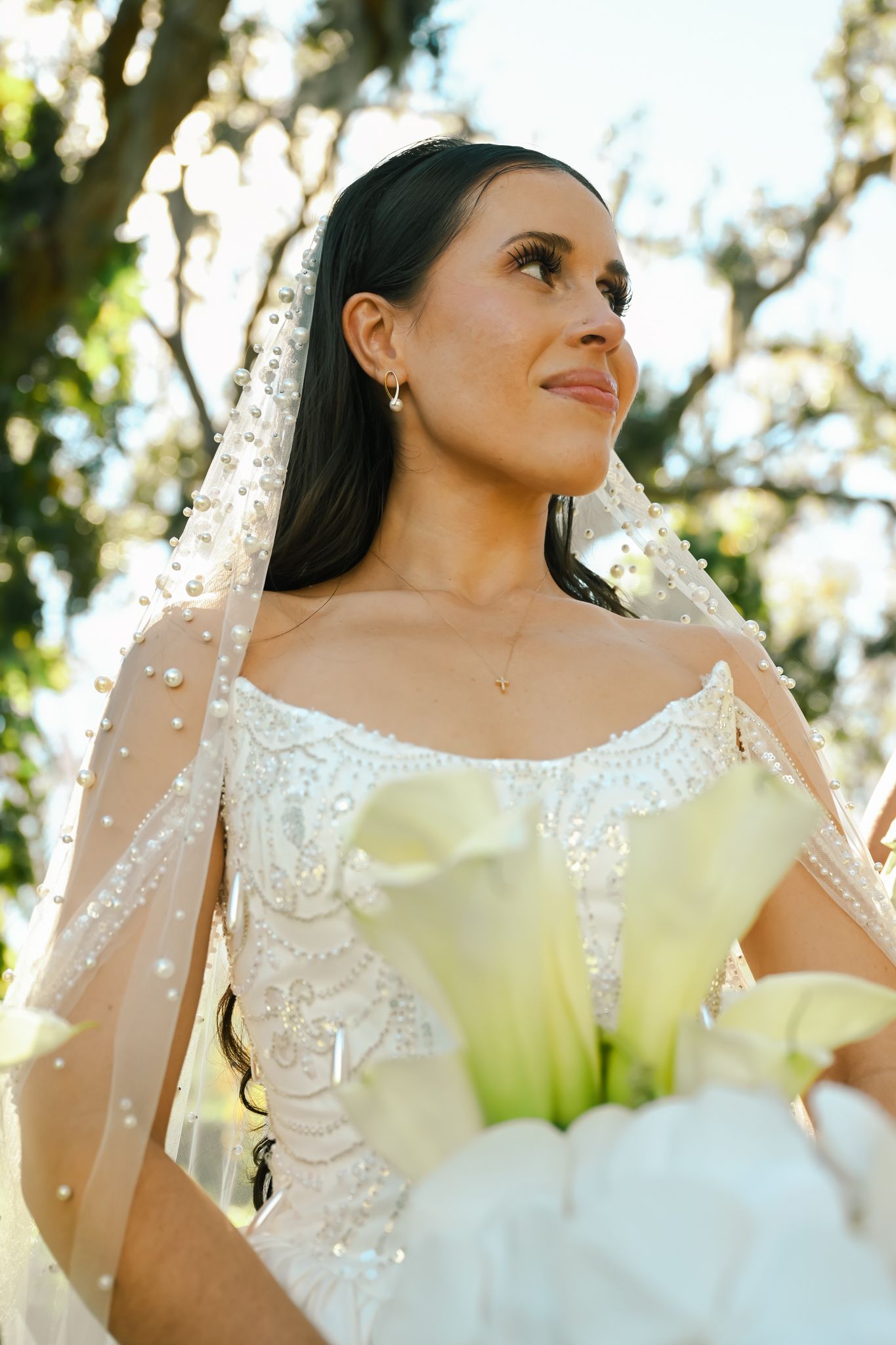 Bride in beaded lace wedding gown and pearl veil holds white calla lily bouquet outdoors under sunlit trees.