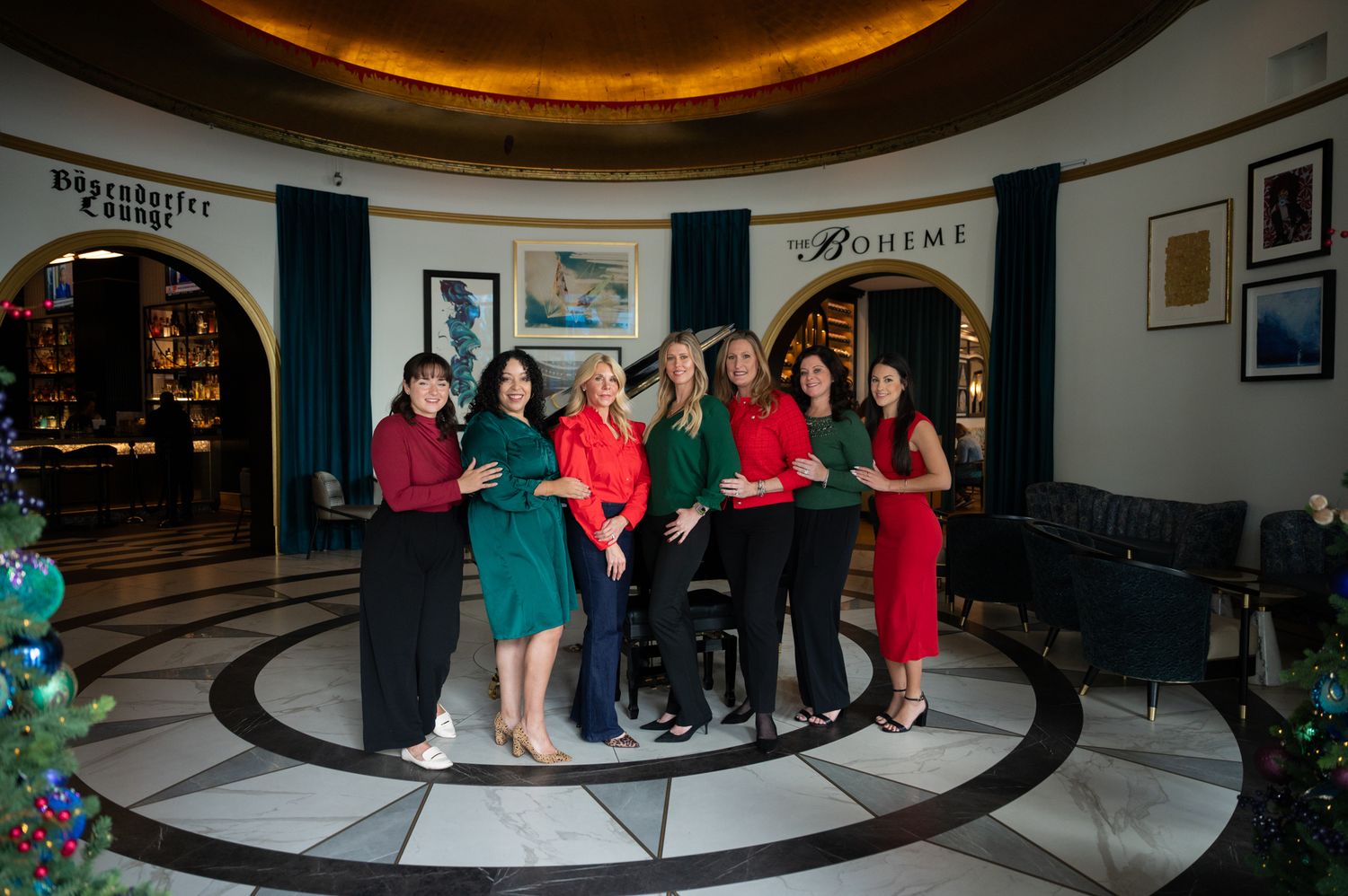 Seven women pose together in an elegant hotel lobby with Boheme restaurant signage and festive holiday decorations.