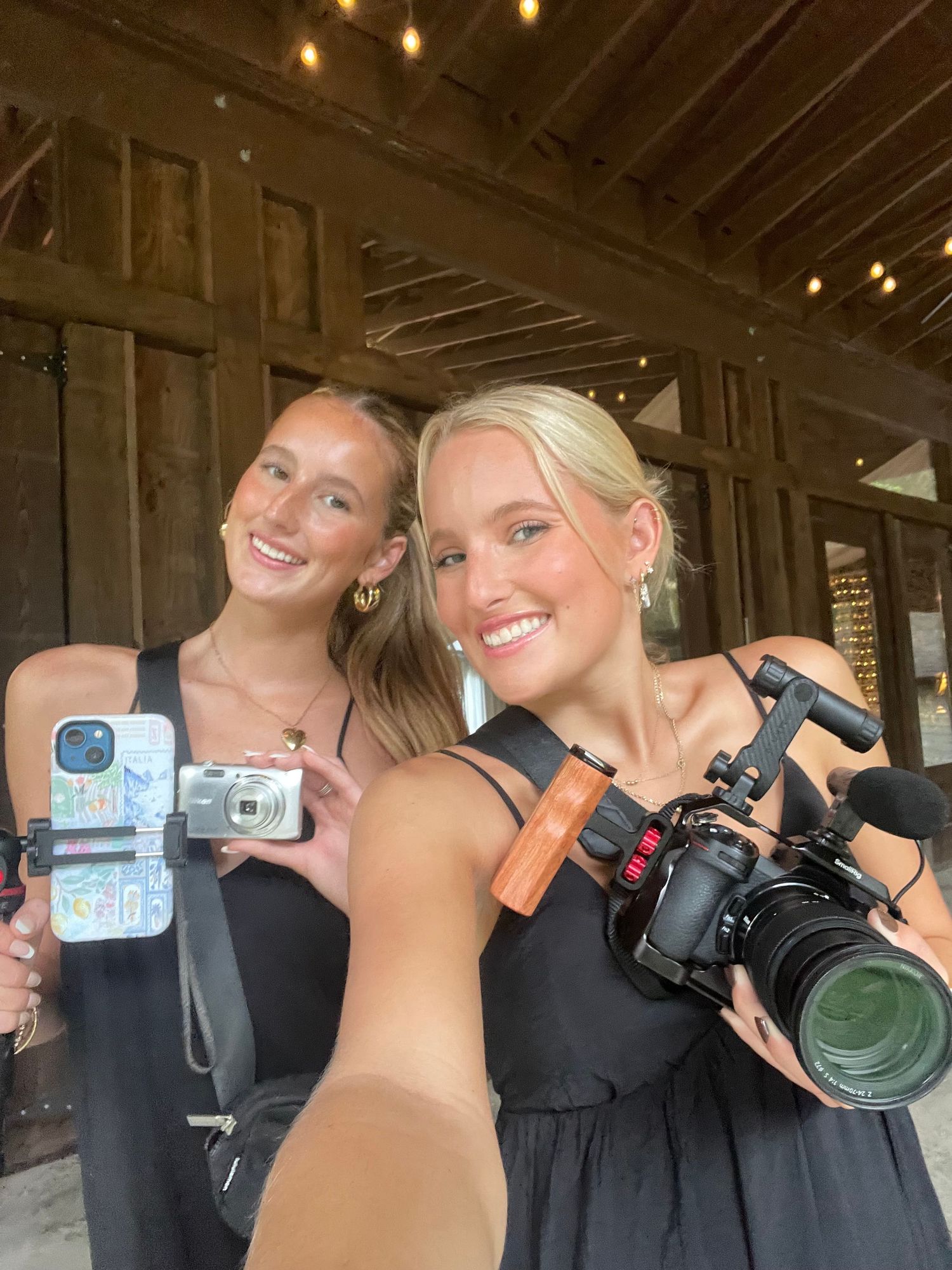 Two smiling blonde women in black dresses pose with cameras including a DSLR and compact camera at an outdoor venue.